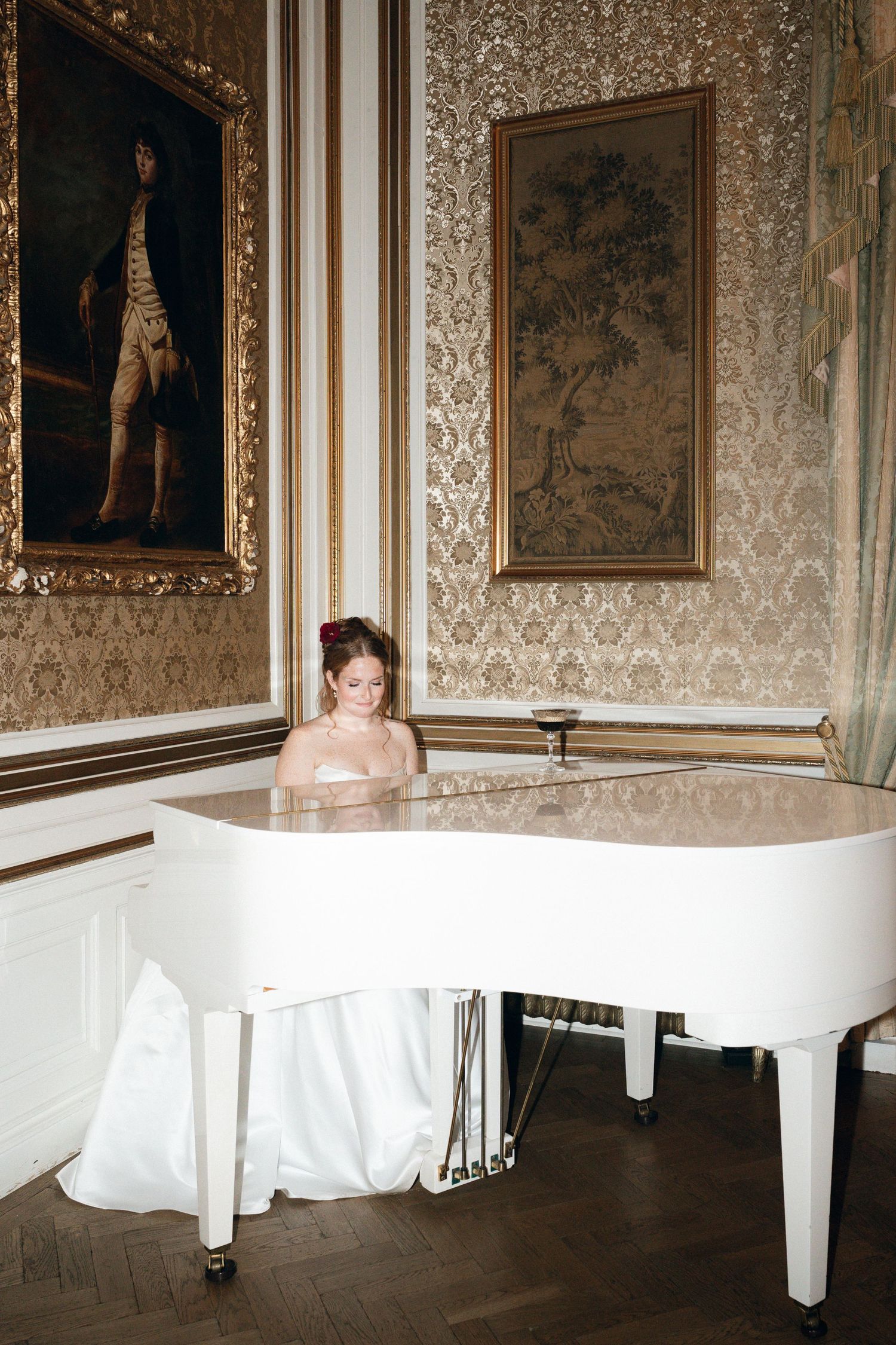A white grand piano sits elegantly in an ornate room with damask wallpaper and gilded framed artwork.