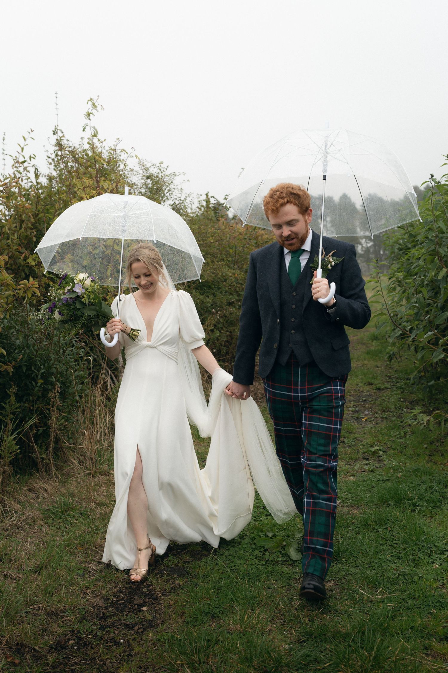 A couple in wedding attire walks through a grassy field holding white umbrellas on a rainy day.