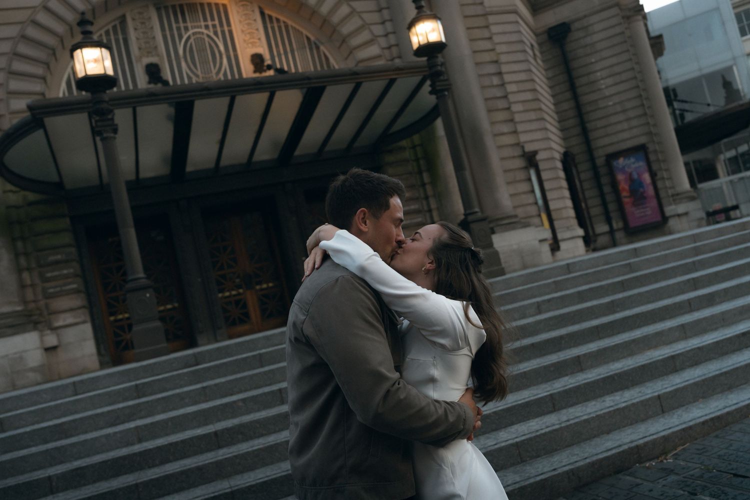 A couple shares a romantic kiss on the steps of a grand building with classical architecture and columns.