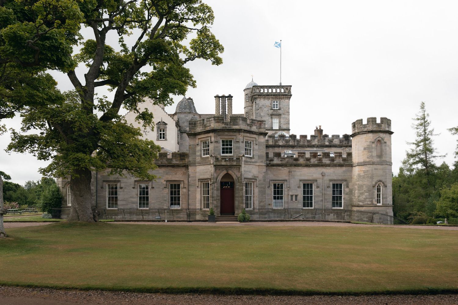 Historic stone castle with turrets and towers stands majestically on a green lawn surrounded by mature trees.