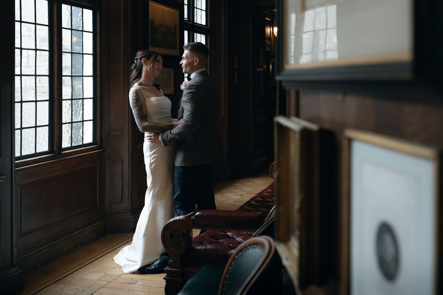 A couple in wedding attire embraces in a dimly lit room with large windows and dramatic shadows.