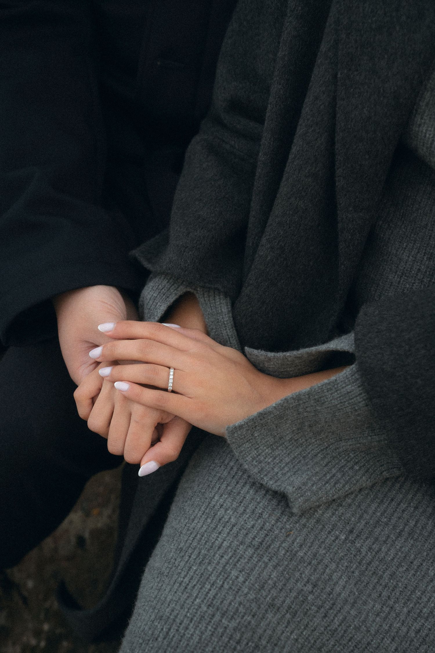 Close-up of hands wearing a delicate silver engagement ring against a dark gray sweater background.