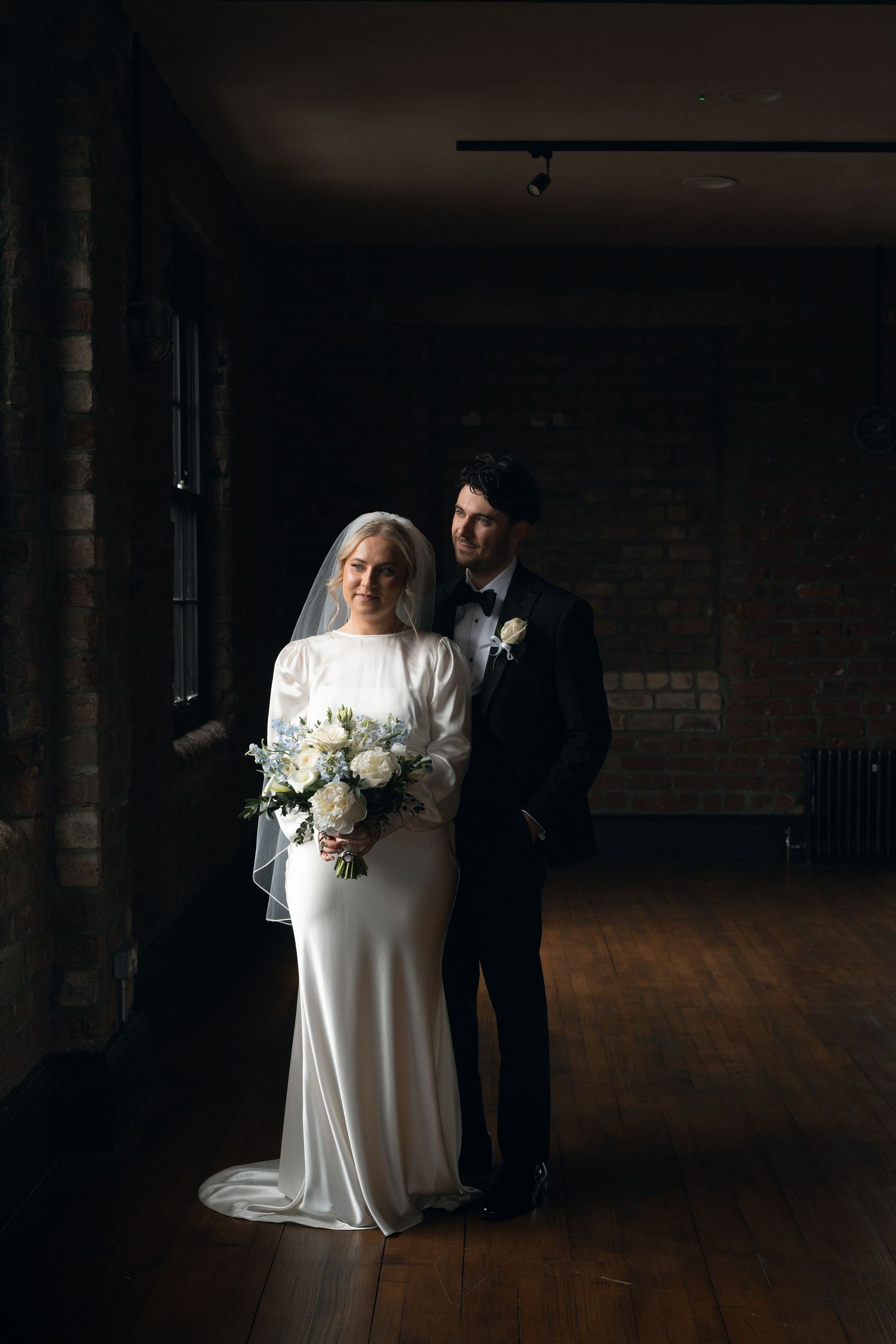 Bride in white gown with veil holds bouquet beside groom in black tuxedo in moody industrial venue.
