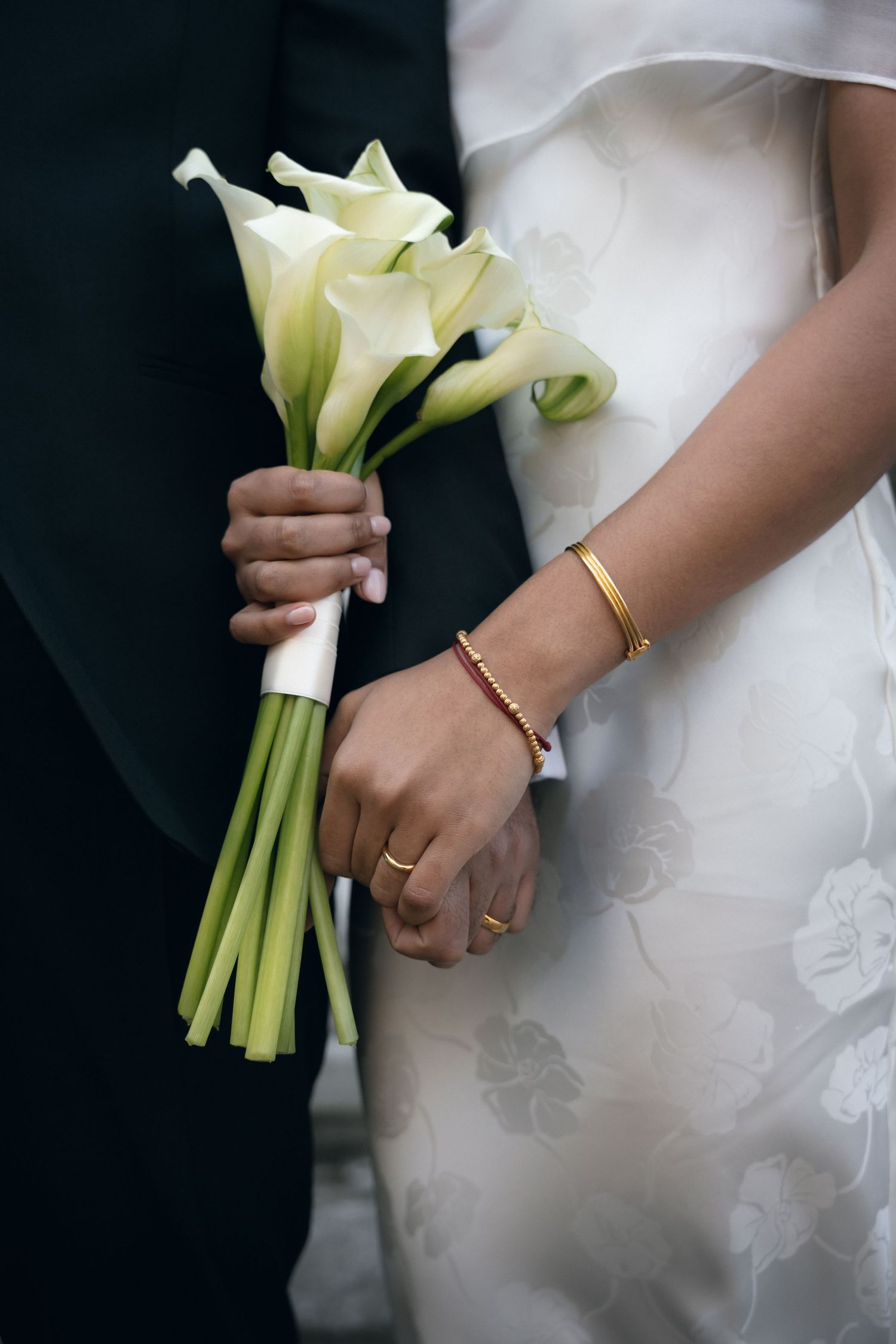 A white calla lily wedding bouquet held against a white wedding dress with delicate floral embroidery details.