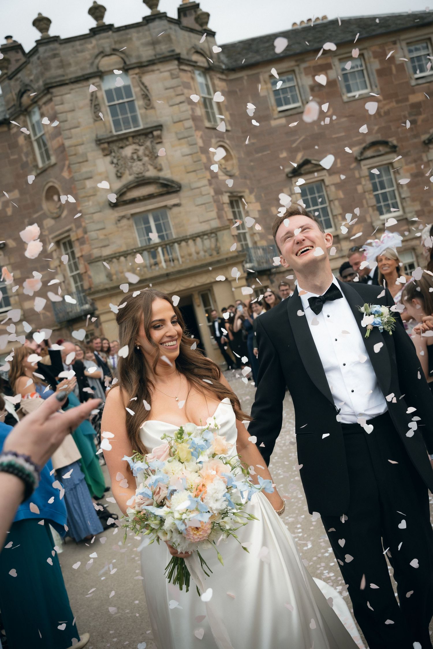A joyful wedding celebration with confetti flying outside a historic stone building as newlyweds walk down the steps.