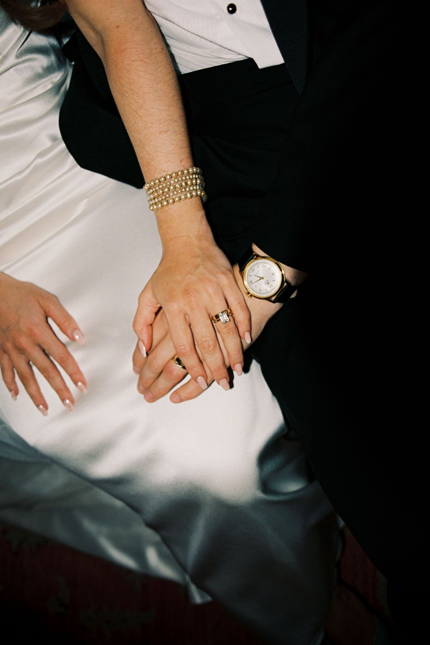 Close-up view of hands wearing gold jewelry resting on white and dark fabric in dramatic lighting.