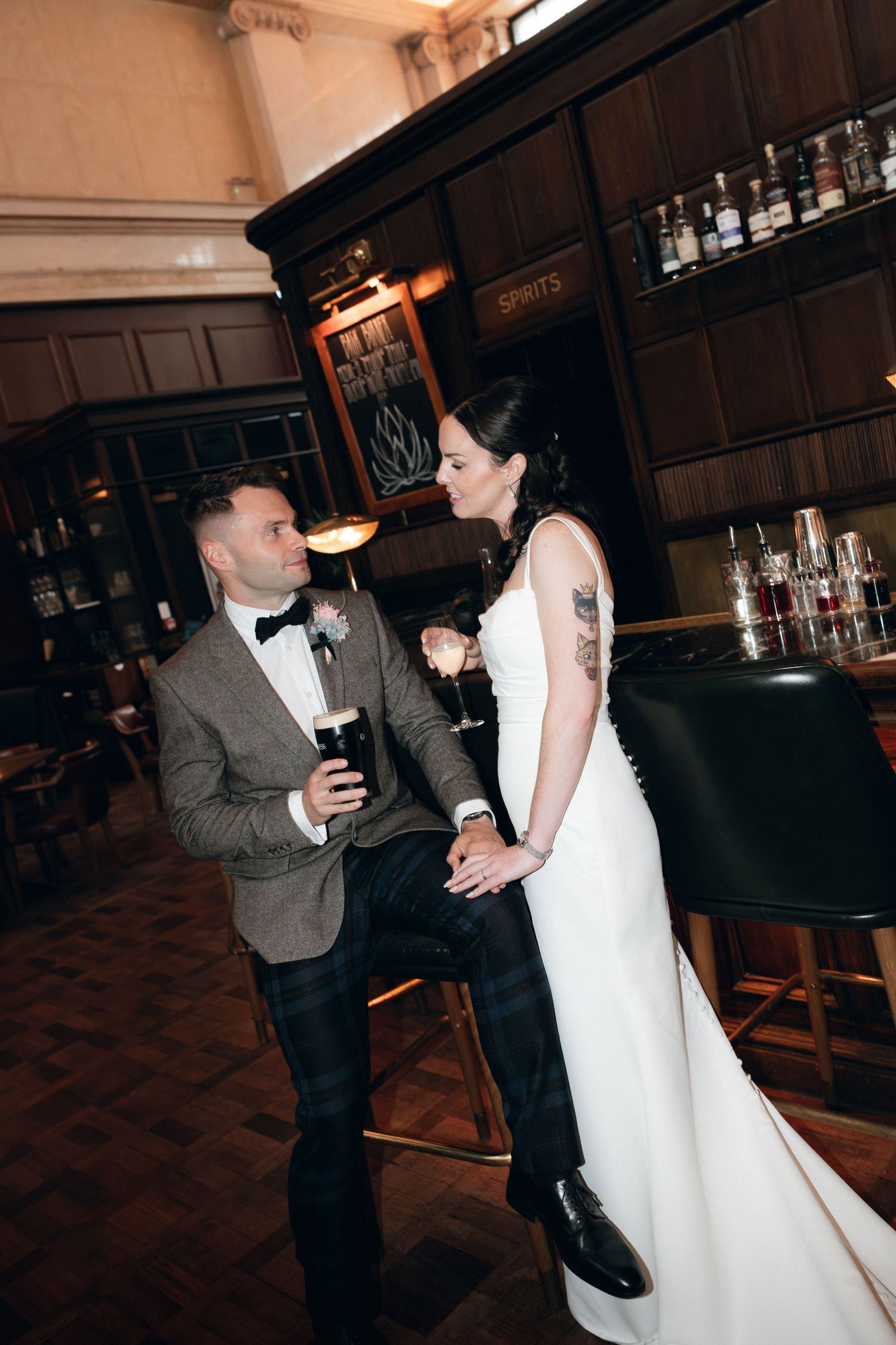 Couple in formal attire share drinks at an elegant bar with dark wood paneling and vintage ambiance.