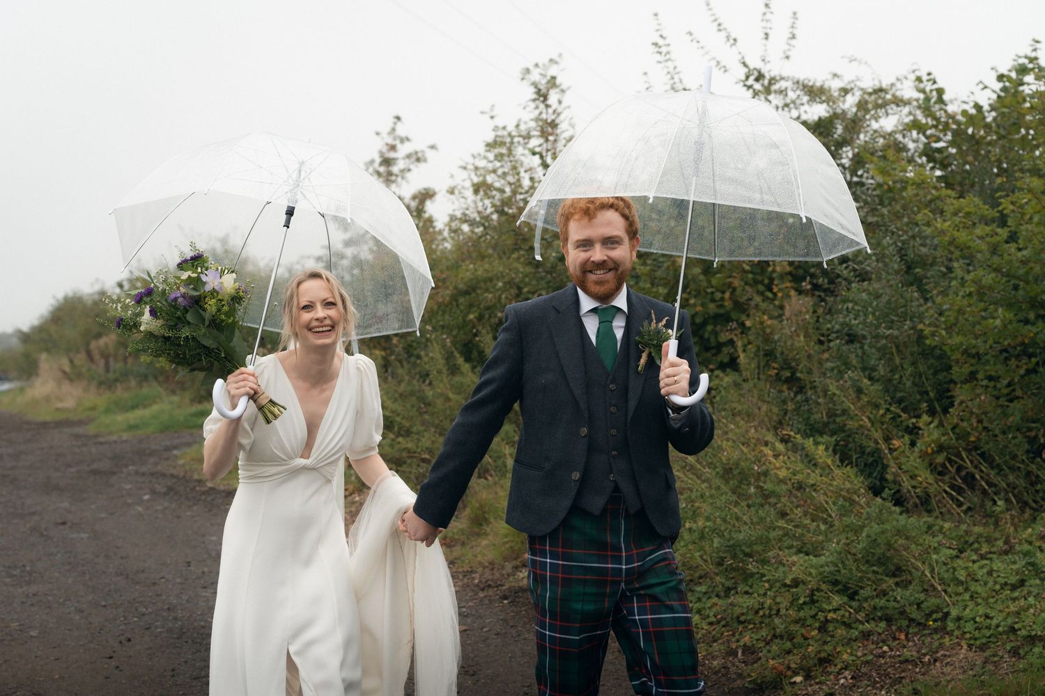 A couple shares a romantic moment under a clear umbrella on their rainy wedding day in a lush outdoor setting.