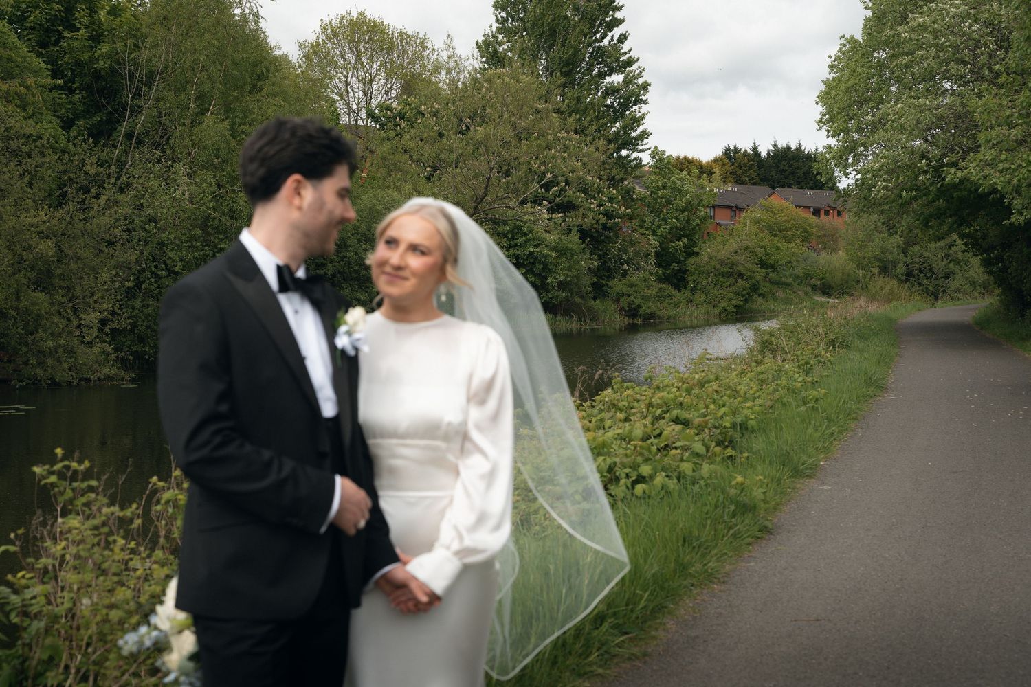 Bride and groom standing by a river path surrounded by lush greenery on their wedding day.