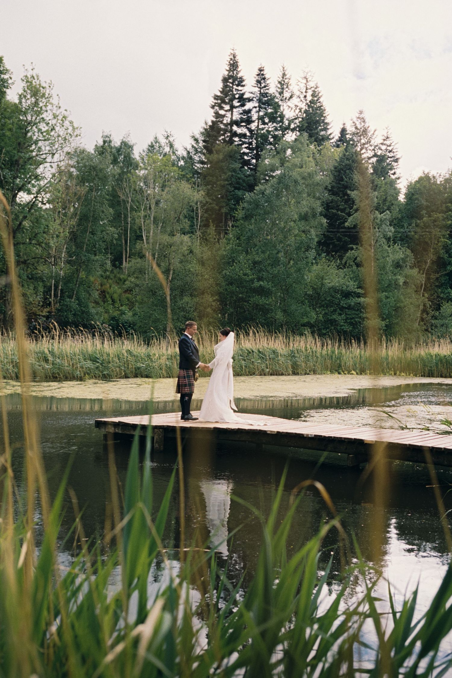 A couple stands on a wooden dock surrounded by evergreen trees and tall grasses at the edge of a tranquil pond.