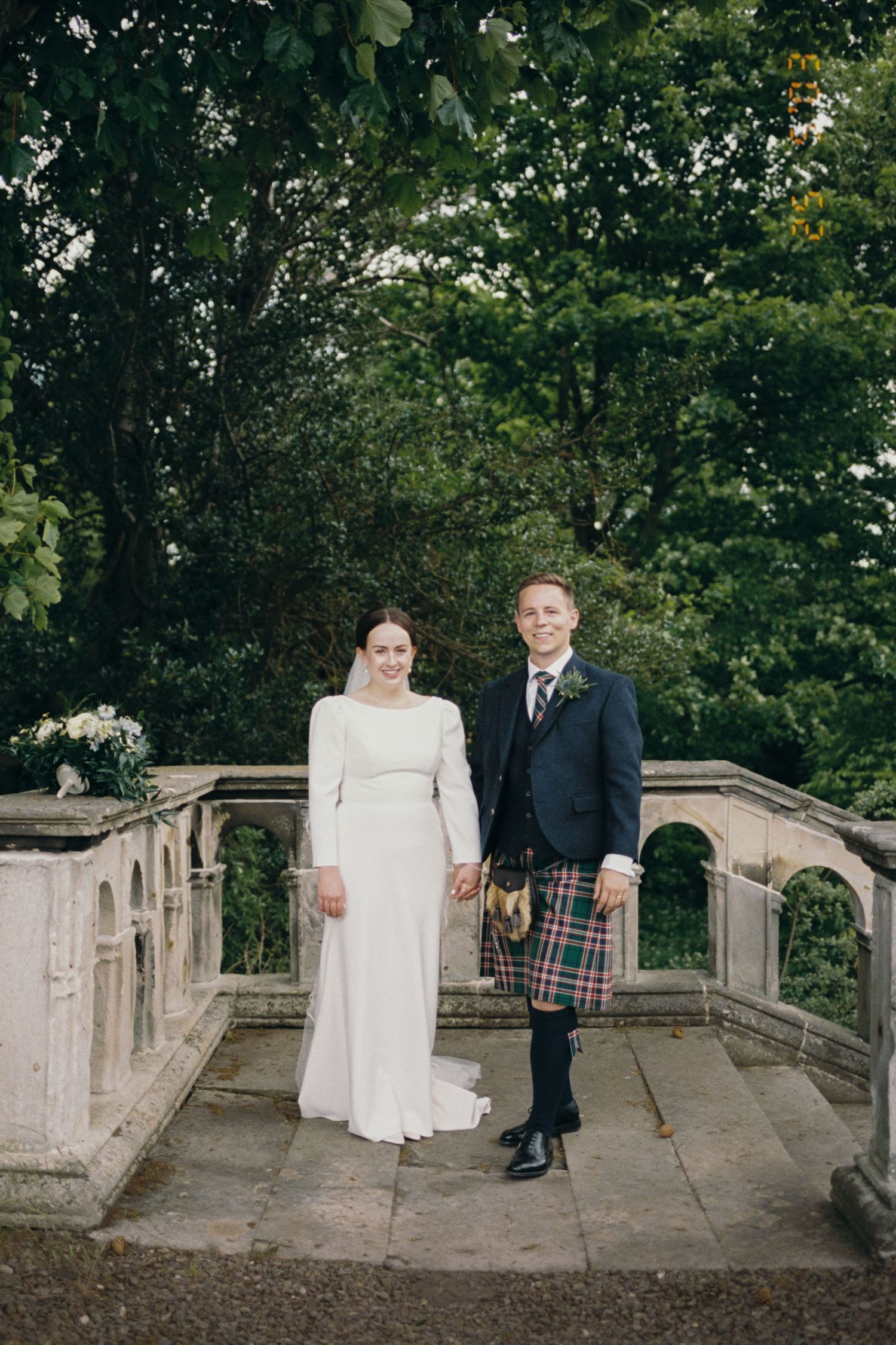 A couple poses on a stone bridge walkway surrounded by lush greenery during their wedding photoshoot.