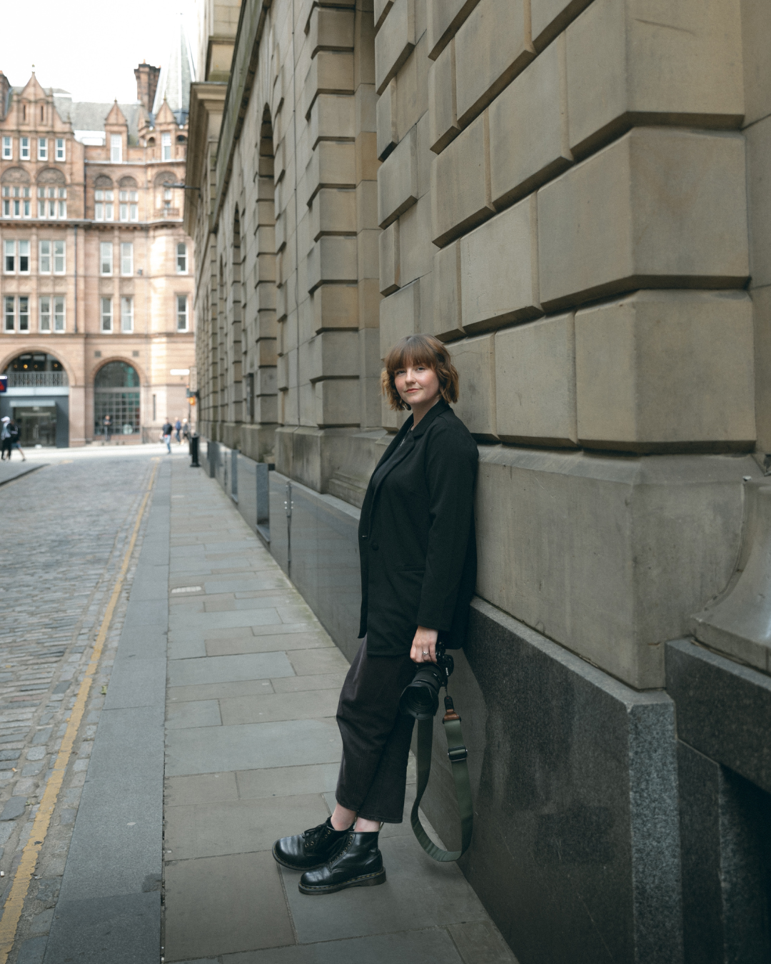 A person in a dark coat and cropped pants poses against a stone building on a historic Edinburgh street.