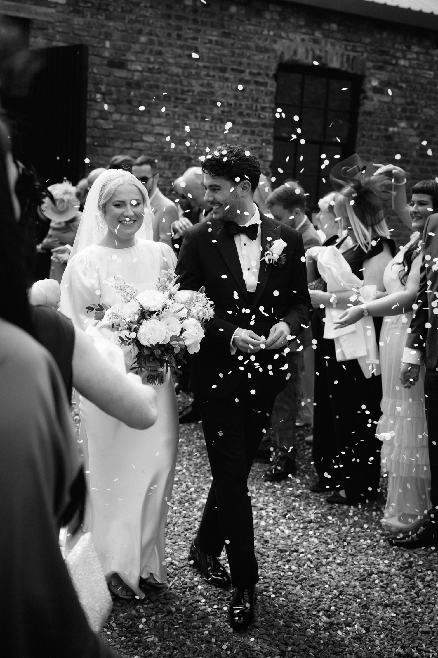 Black and white photo of a bride and groom walking through confetti shower surrounded by cheering wedding guests.
