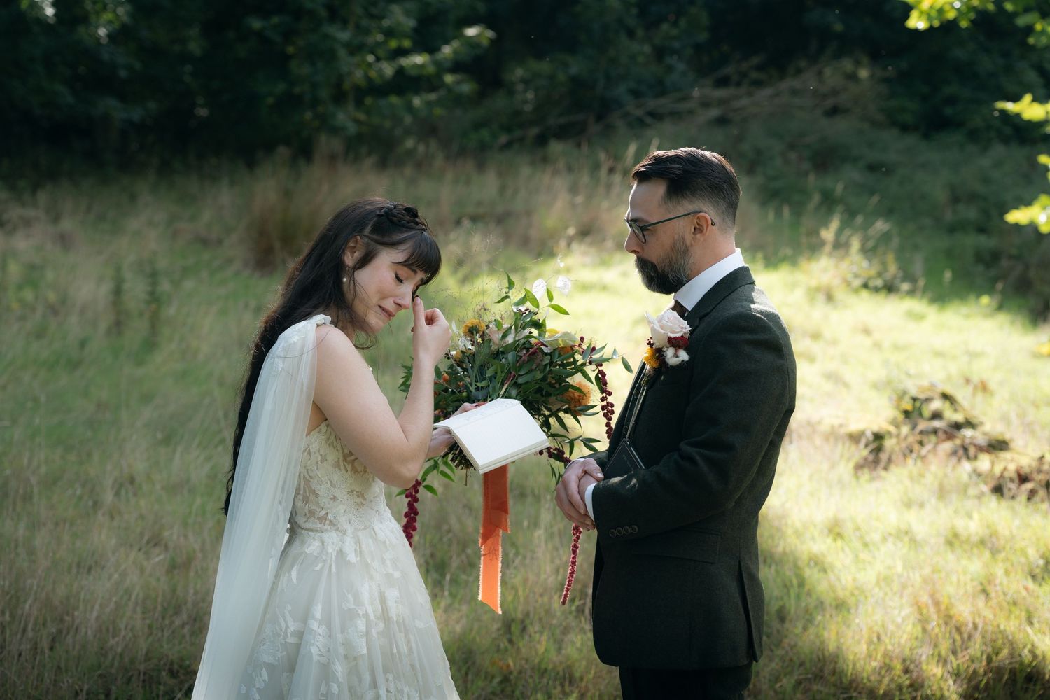 A couple in wedding attire shares an intimate moment outdoors during their wedding ceremony in a sunlit meadow.