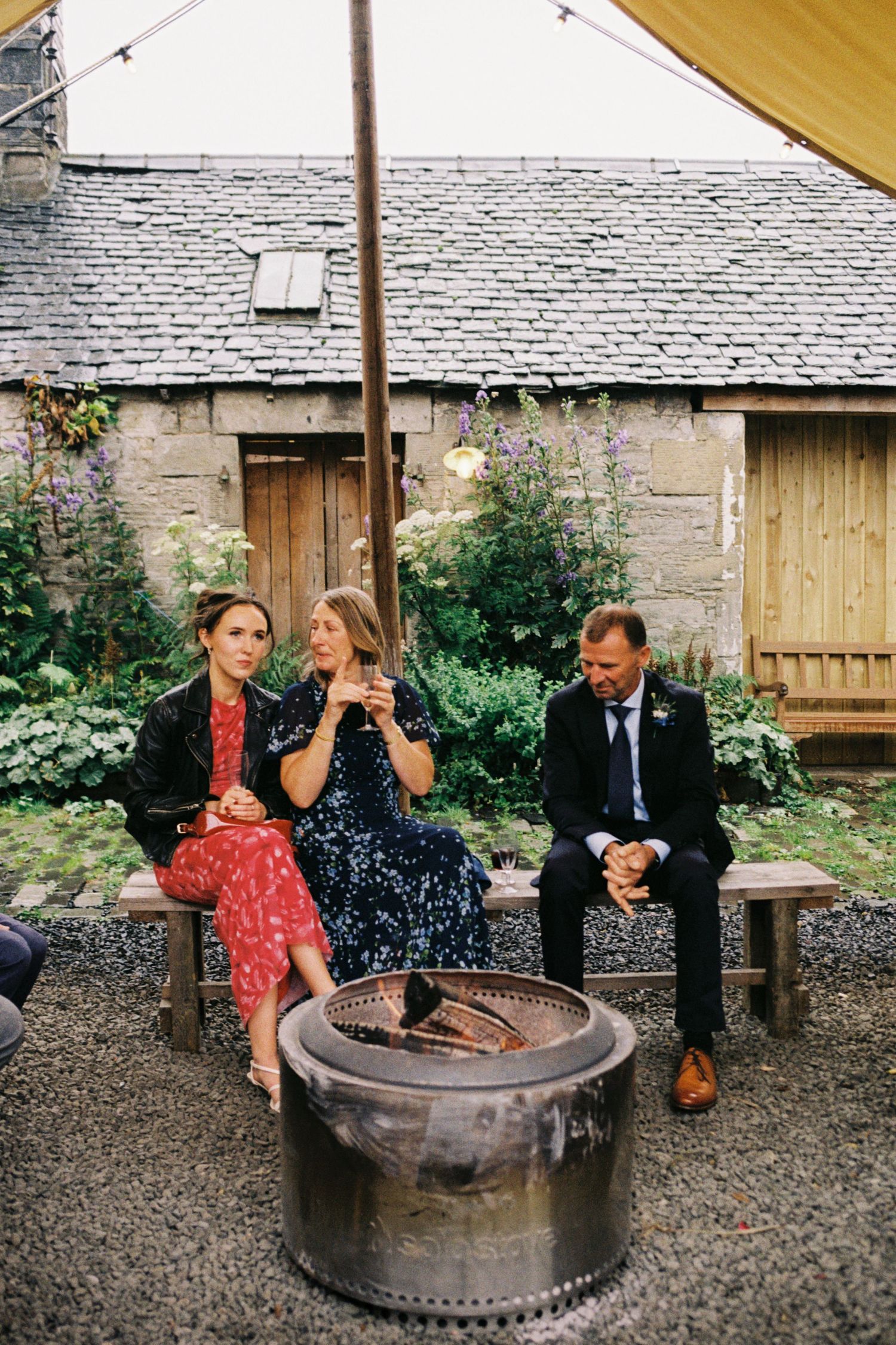 Three people sit and chat on a rustic tire swing in a garden setting with stone walls and wooden doors.
