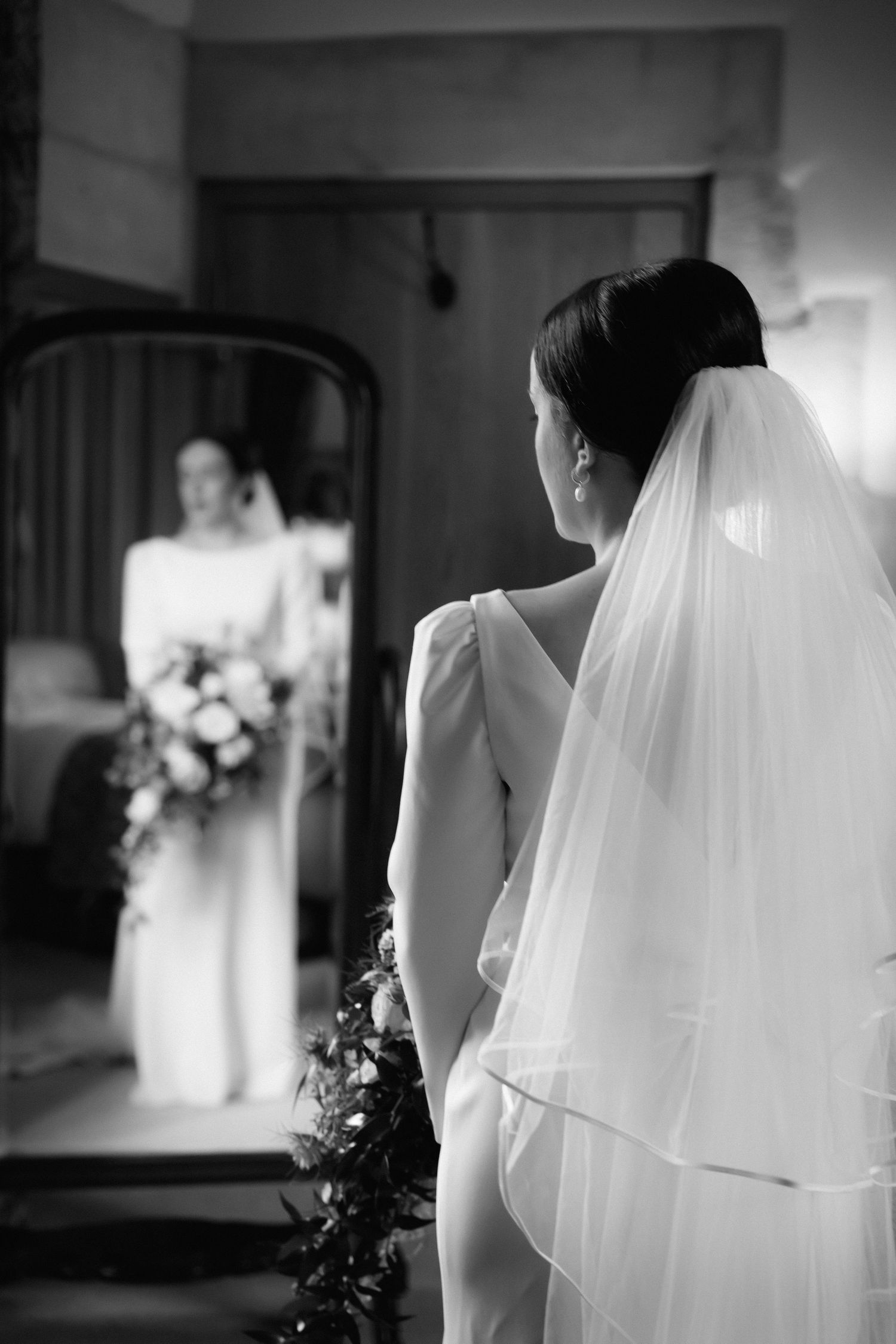 A bride in a white wedding dress and flowing veil looks at her reflection in a mirror during a black and white photo.