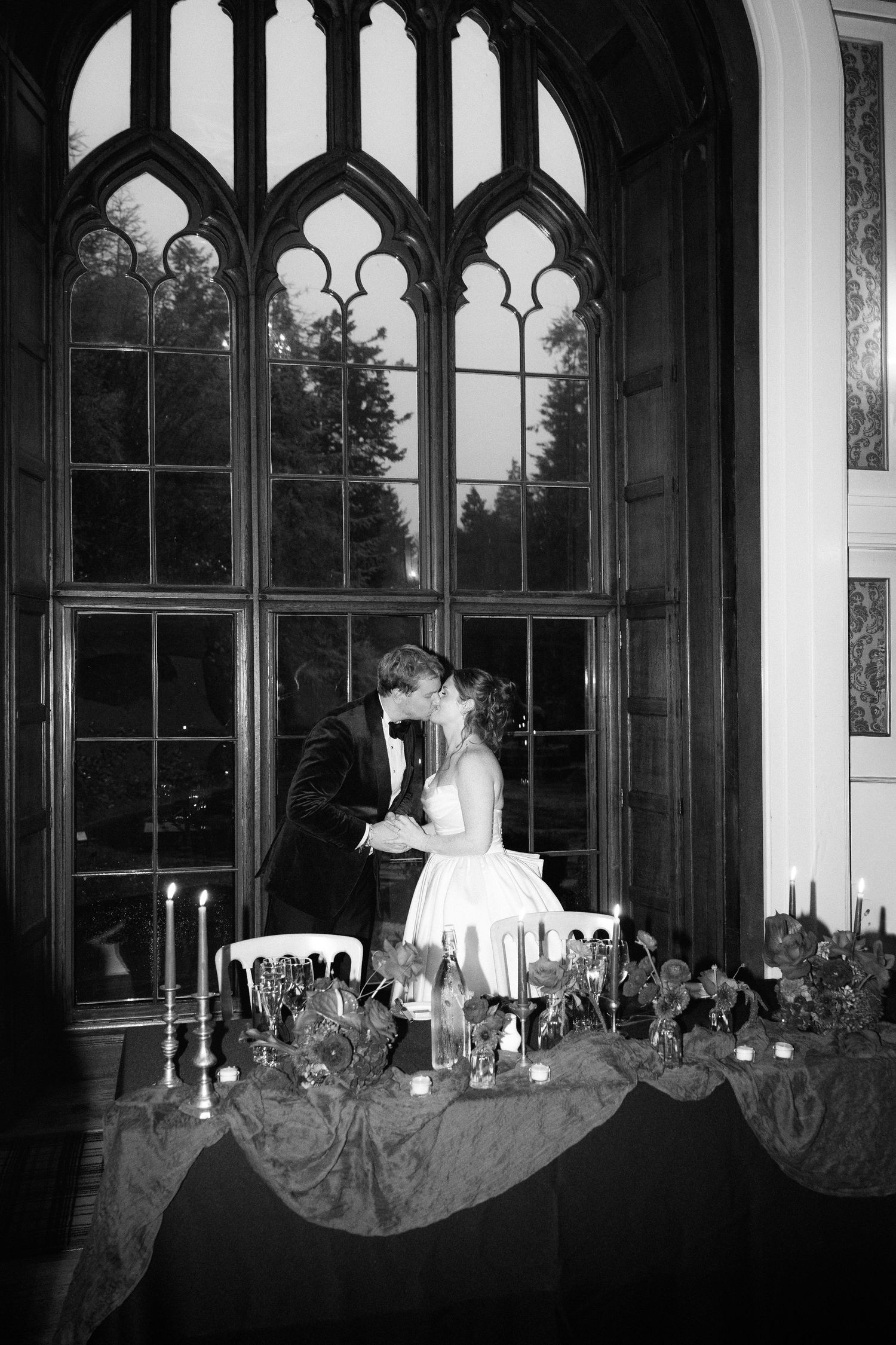 A romantic black and white wedding scene with a couple silhouetted against a Gothic arched window with candlelit decor.