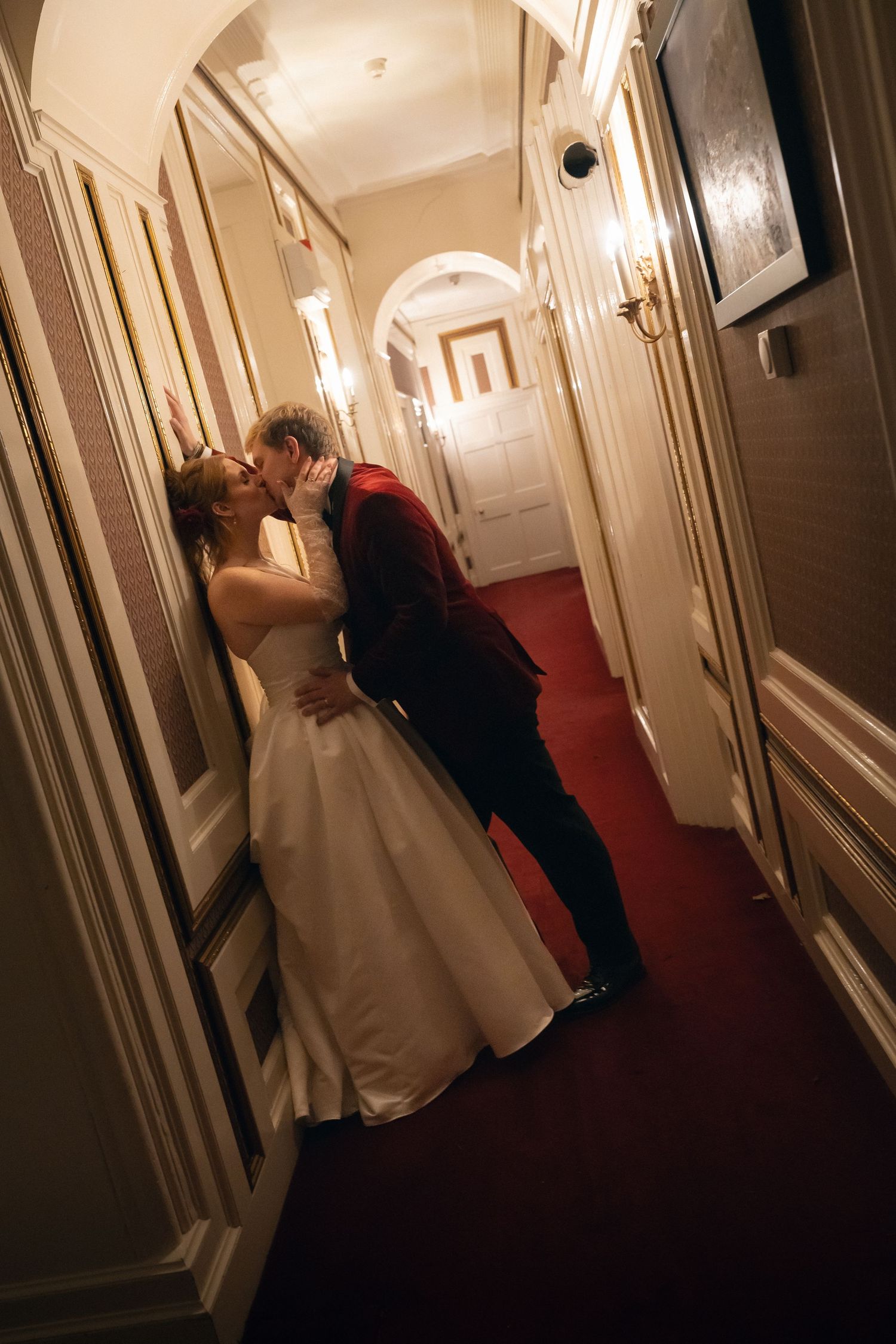 A couple shares a romantic moment in a dimly lit hallway with cream colored walls and framed artwork.