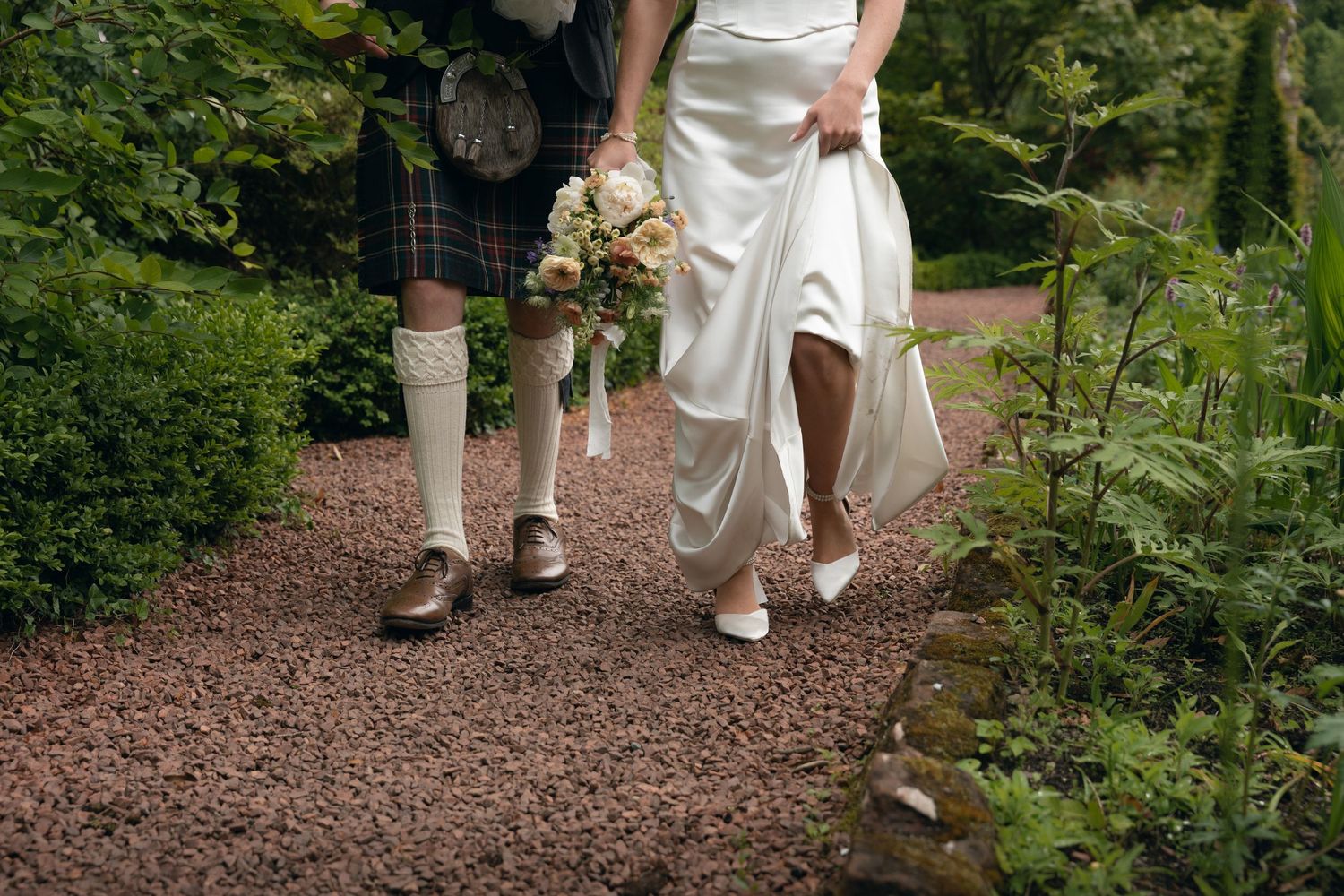 A pair walking on a garden path shows traditional Scottish kilt and white wedding dress with bouquet in hand.
