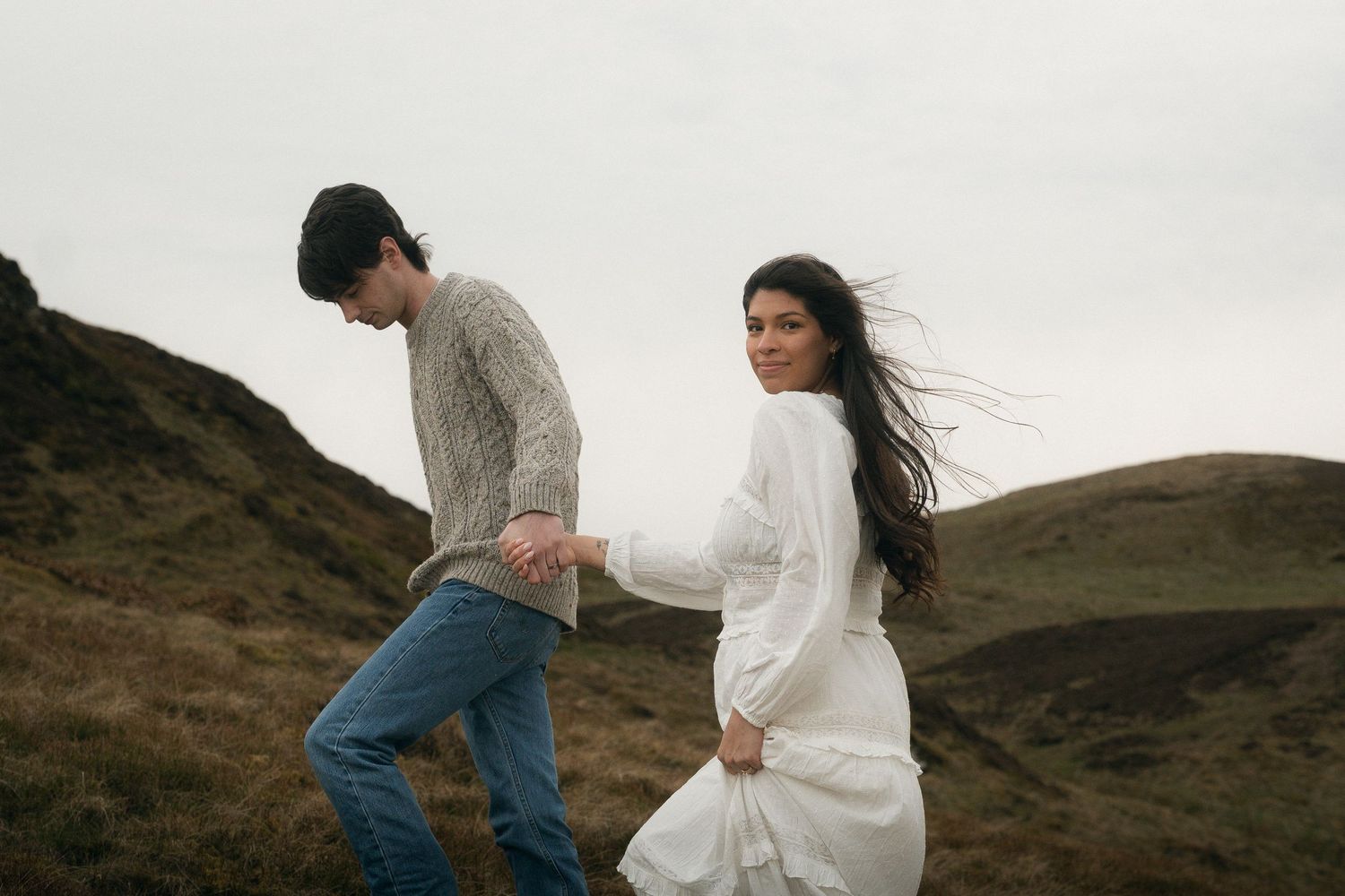 Couple holding hands on misty highland hills, woman in white dress with wind-blown hair glancing back romantically.