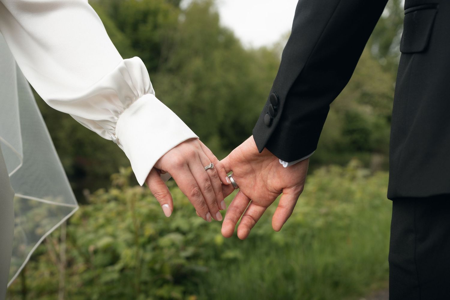 Bride and groom holding hands outdoors, showing wedding rings, wearing white dress and black suit.