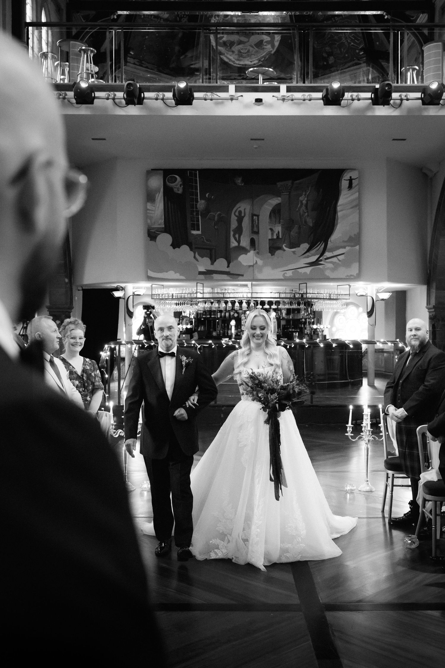 Black and white photo of a bride walking down the aisle with her father at an elegant indoor wedding ceremony.