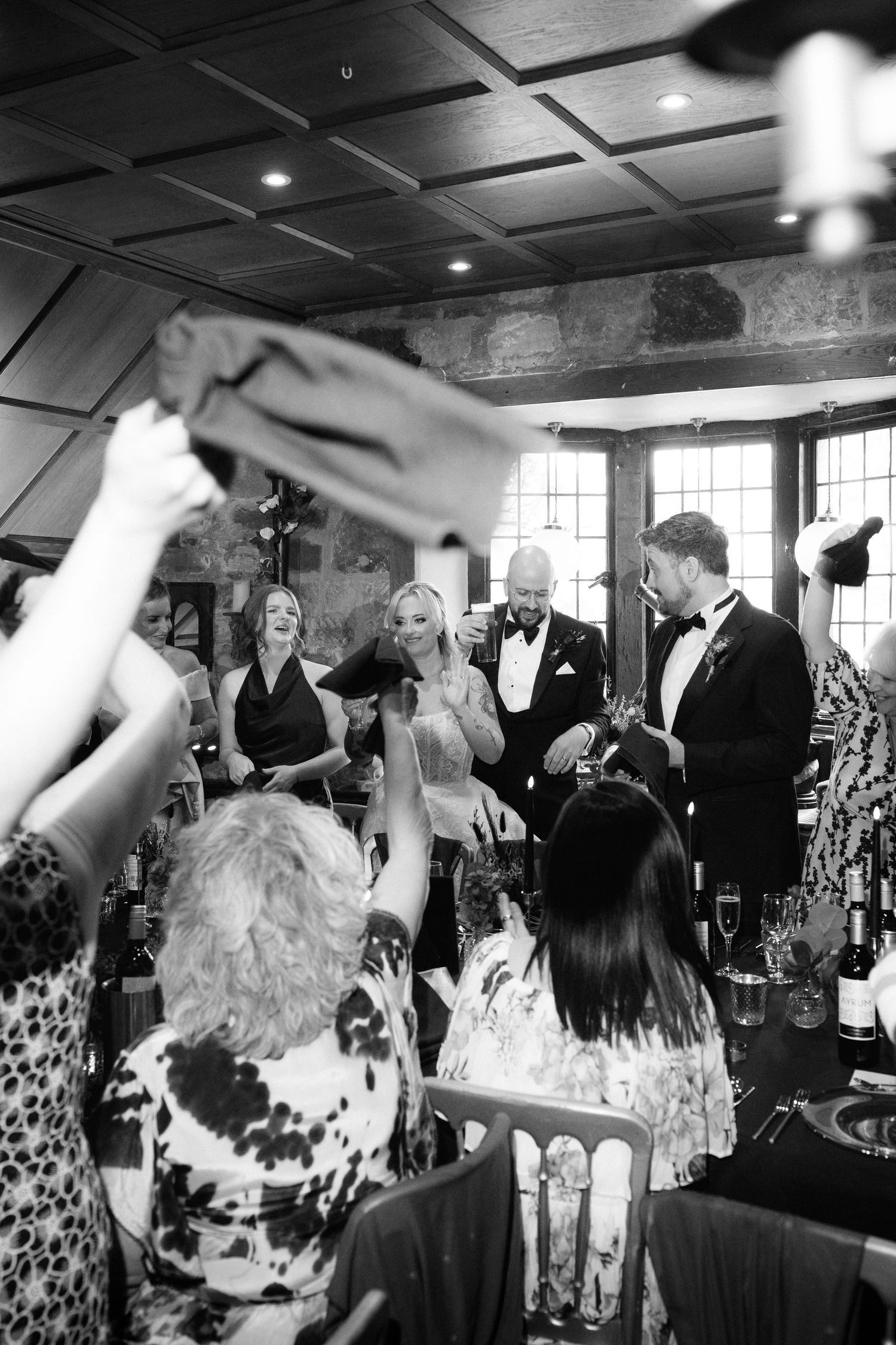Black and white wedding reception photo with guests raising glasses in a celebratory toast inside an elegant venue.
