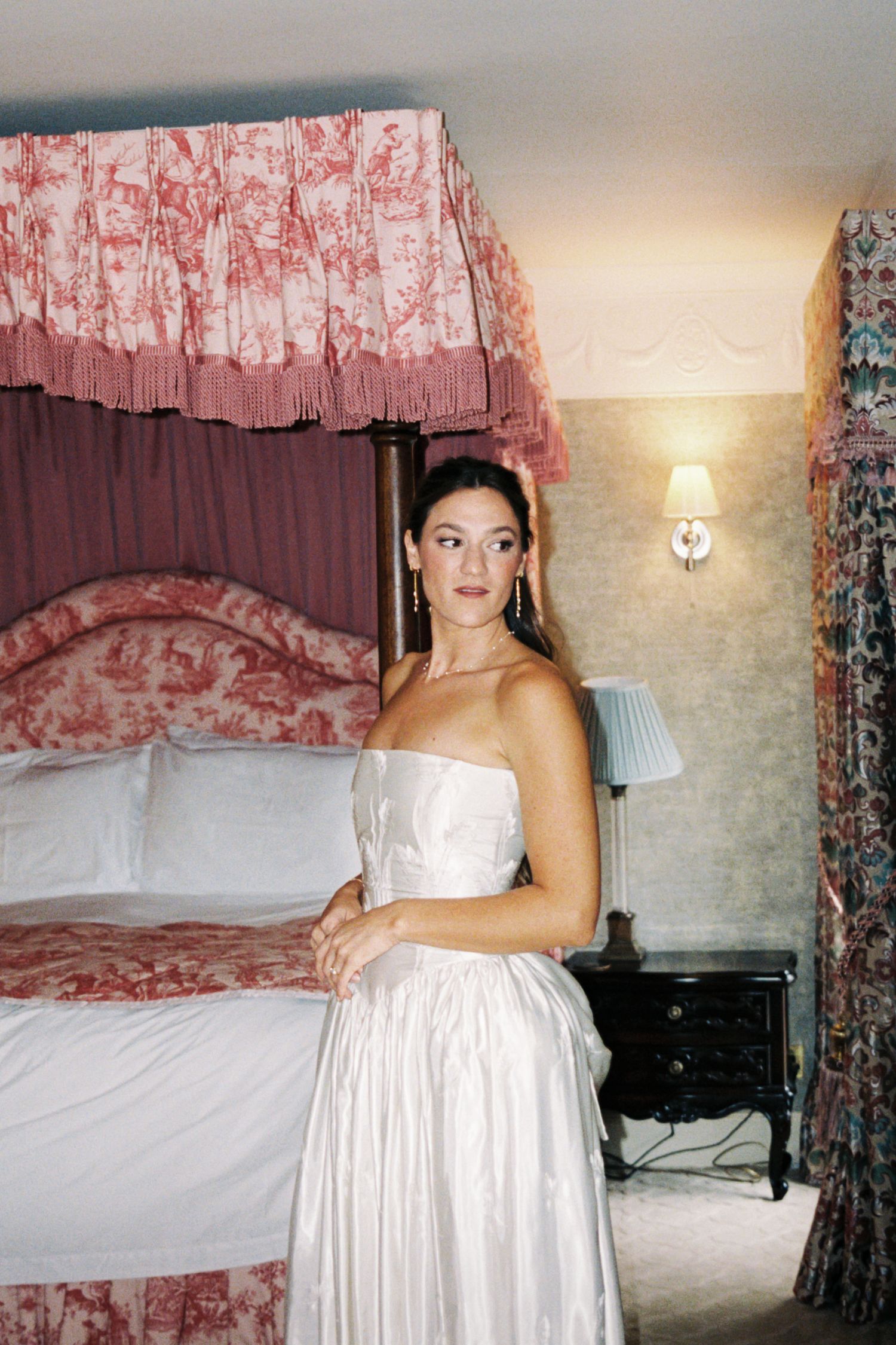 A bedroom interior with red and white toile curtains and bedding featuring a white dress on display.