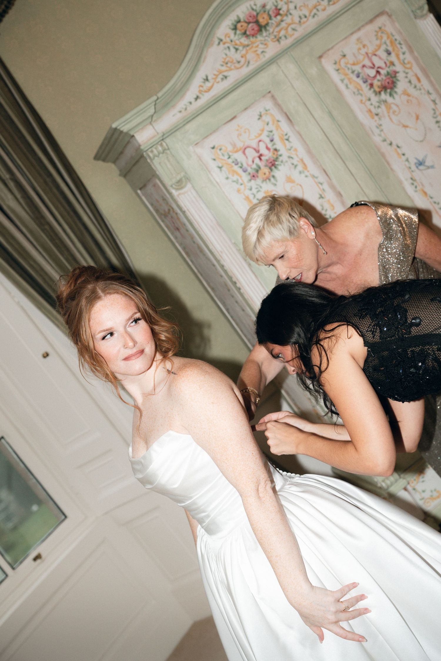 A bride in a white dress getting help with final adjustments in an ornate room with decorative wall panels.