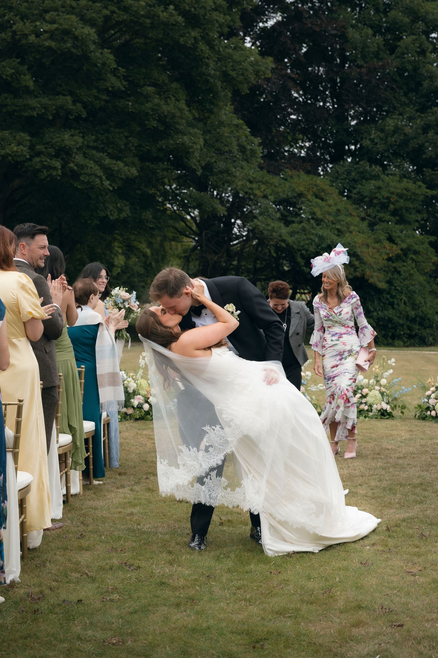 Wedding party members in formal attire playfully dip and pose together on a grassy lawn during outdoor photo session.