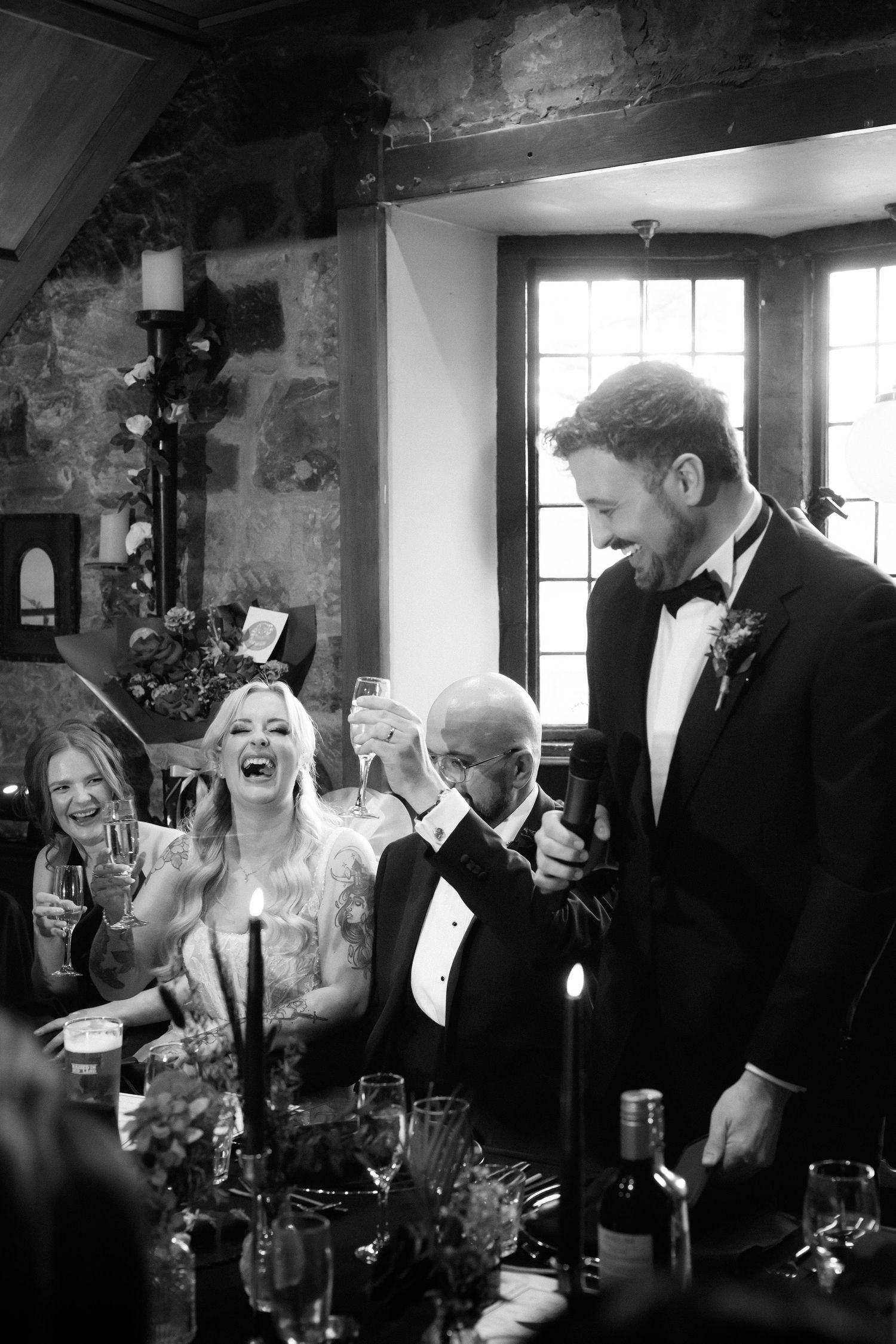 Black and white wedding reception toast with laughing bride in gown and groom in tuxedo raising glasses in rustic stone venue.