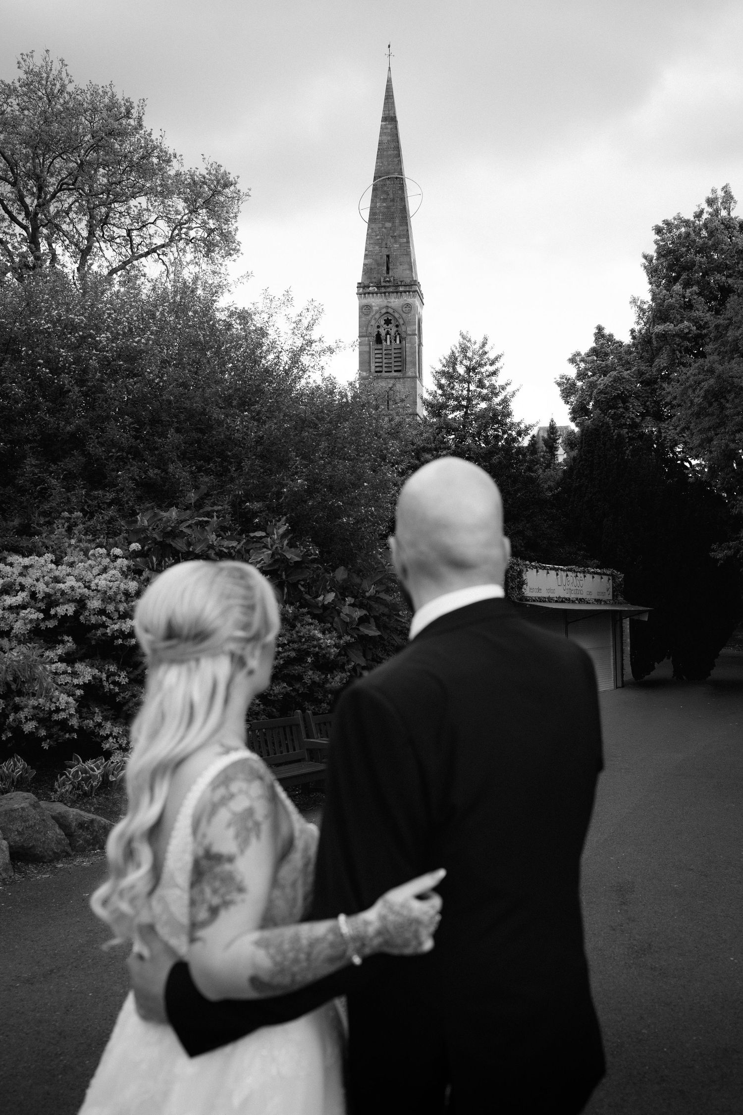 Black and white photo of tattooed bride and groom walking toward a church steeple surrounded by lush trees.