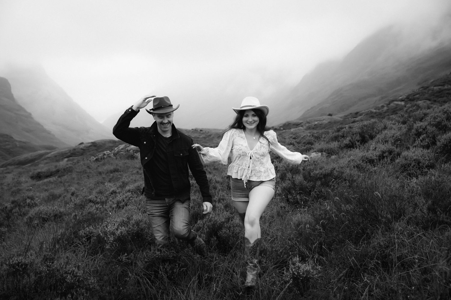 Black and white panoramic sequence showing two people dancing joyfully on a misty mountainside slope.