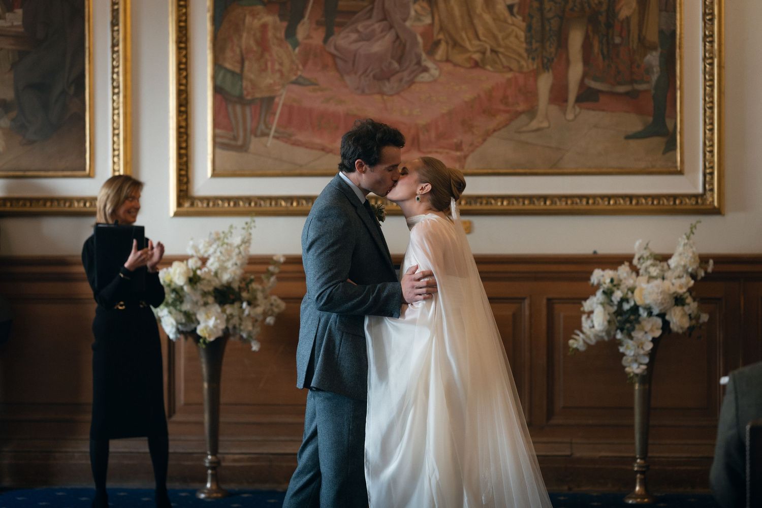 A romantic wedding ceremony in an elegant room with ornate wall paintings and floral arrangements on pedestals.