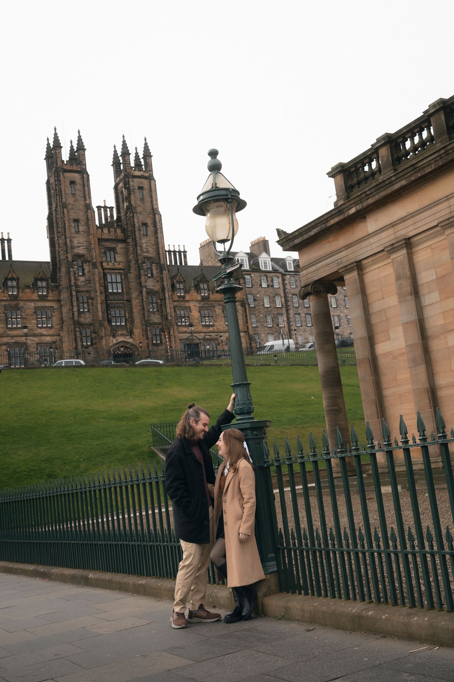 Couple standing by a vintage street lamp near historic Edinburgh building with Gothic architecture and green iron fence.