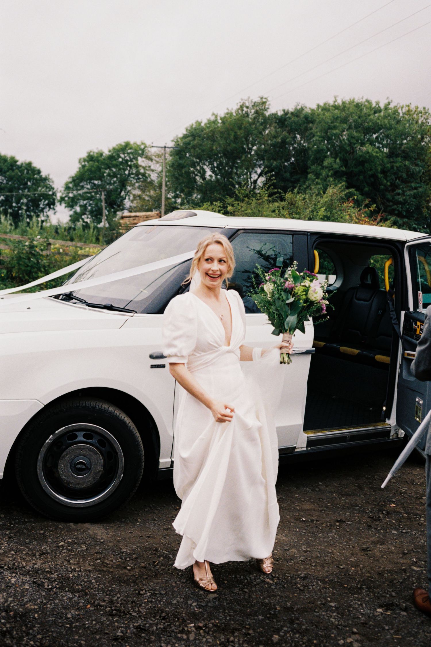 A classic white London taxi parked outdoors with someone in a flowing white dress holding a floral bouquet.
