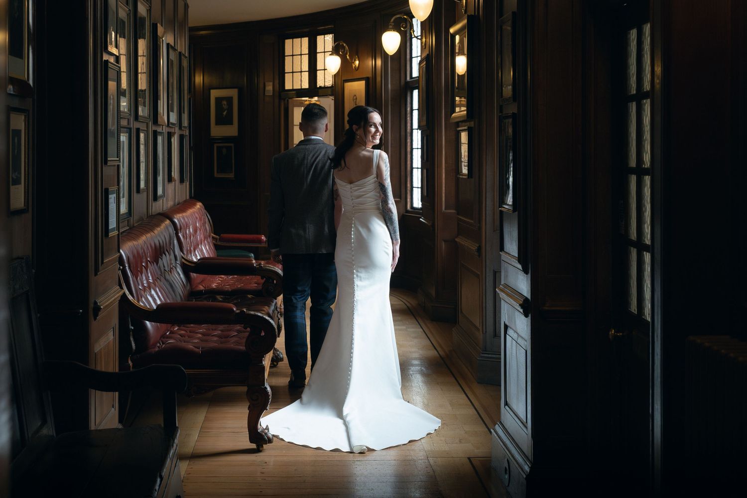 A bride in a white wedding dress stands in a dark hallway with elegant lighting and framed artwork on the walls.