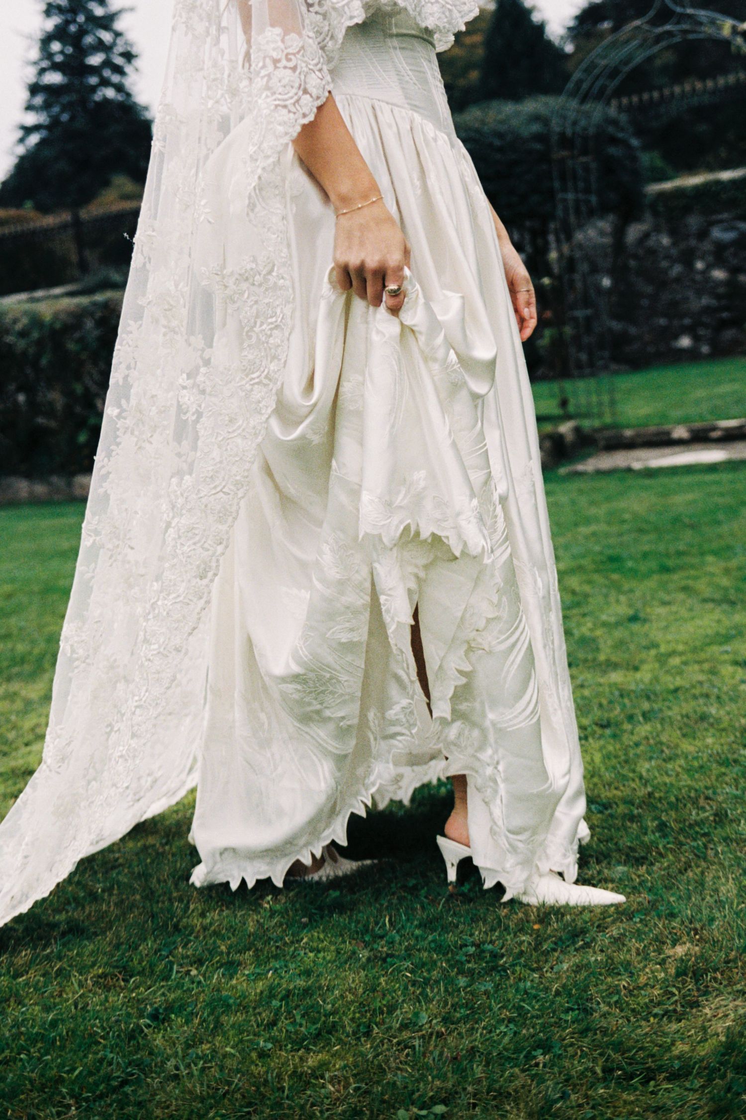 Close-up details of a white lace wedding dress flowing over green grass outdoors.
