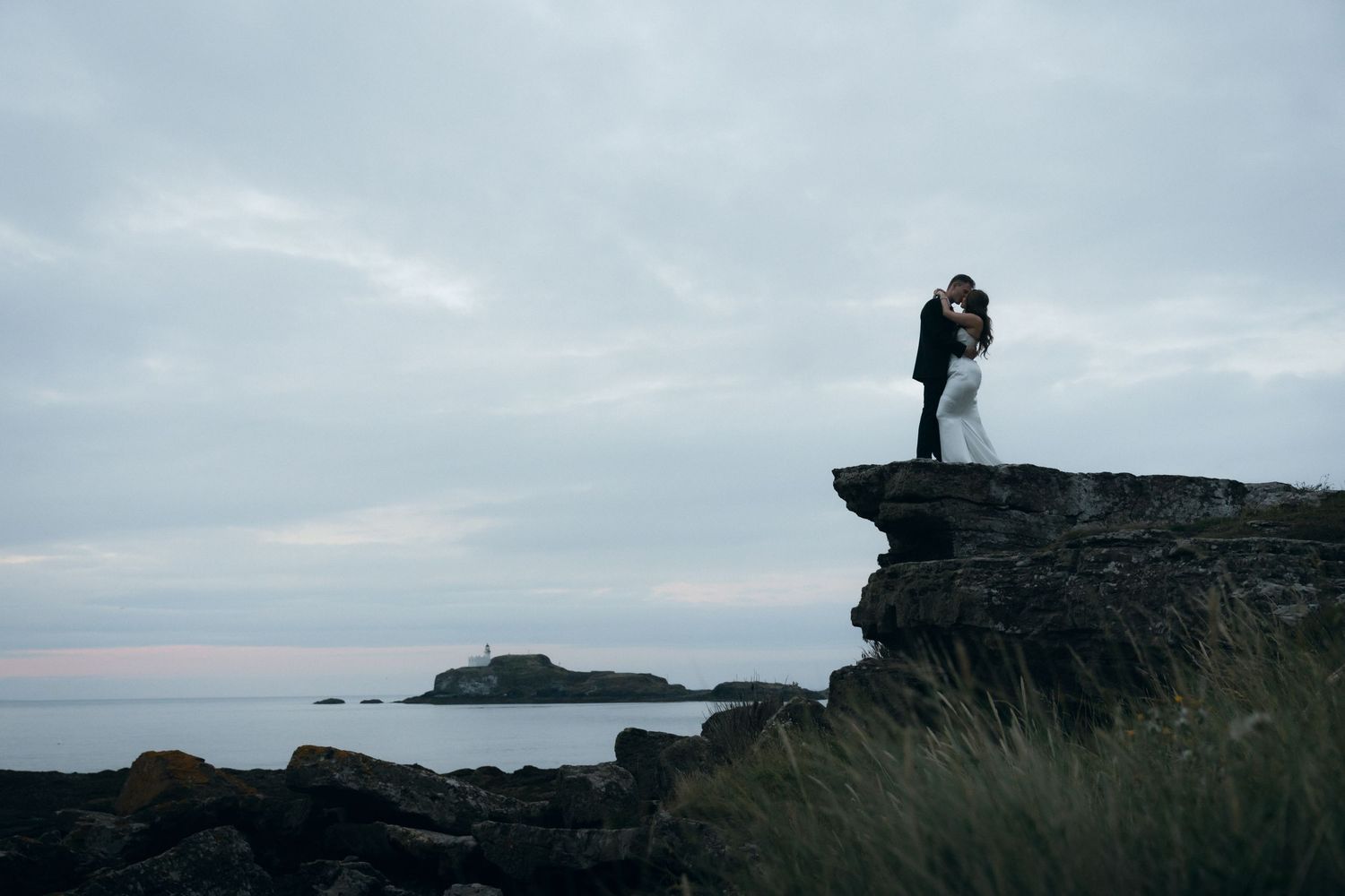 A couple stands silhouetted against a dramatic coastal landscape during their wedding photoshoot at sunset.