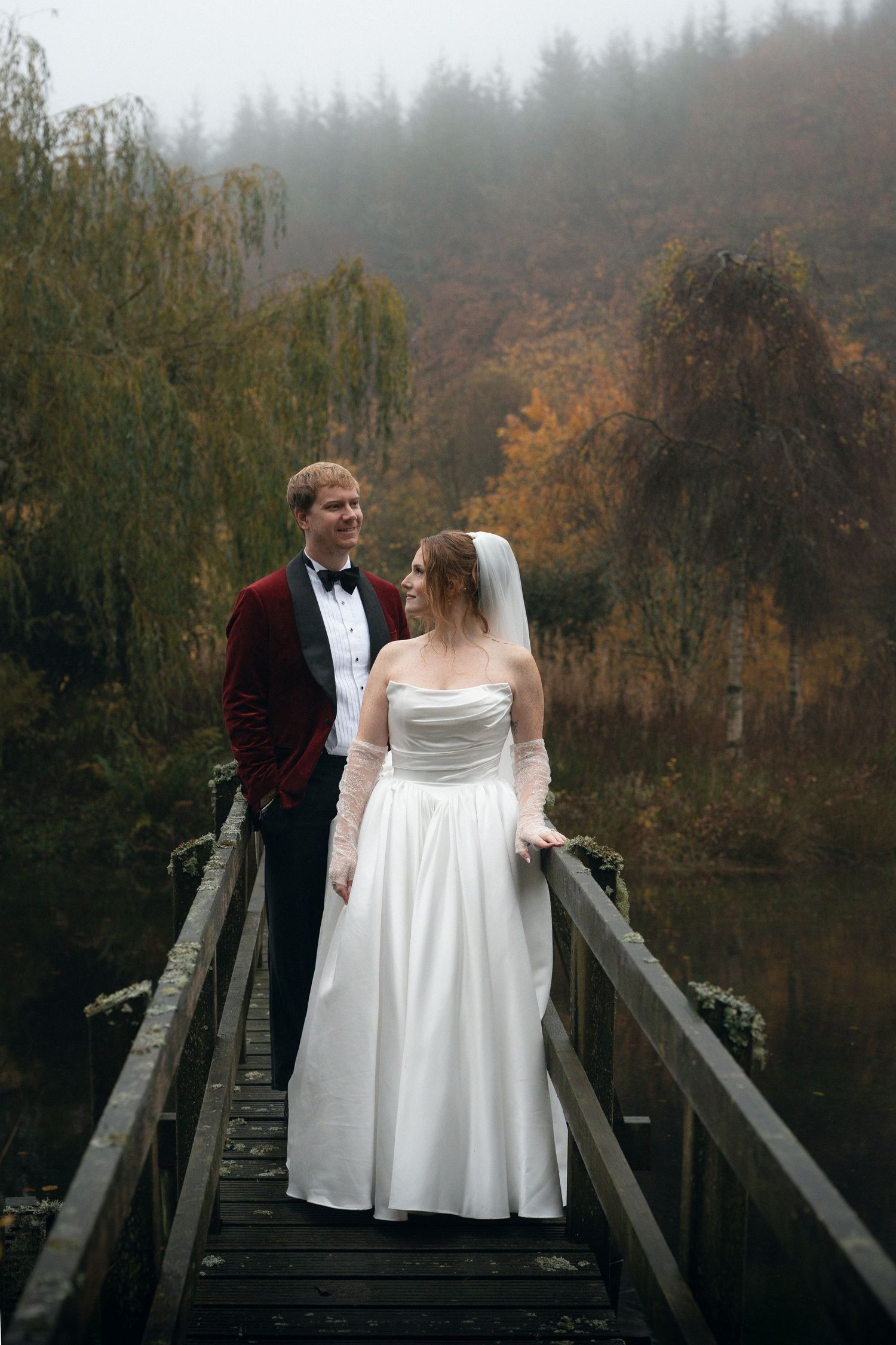 A couple in wedding attire stands together on a wooden bridge surrounded by misty autumn trees.