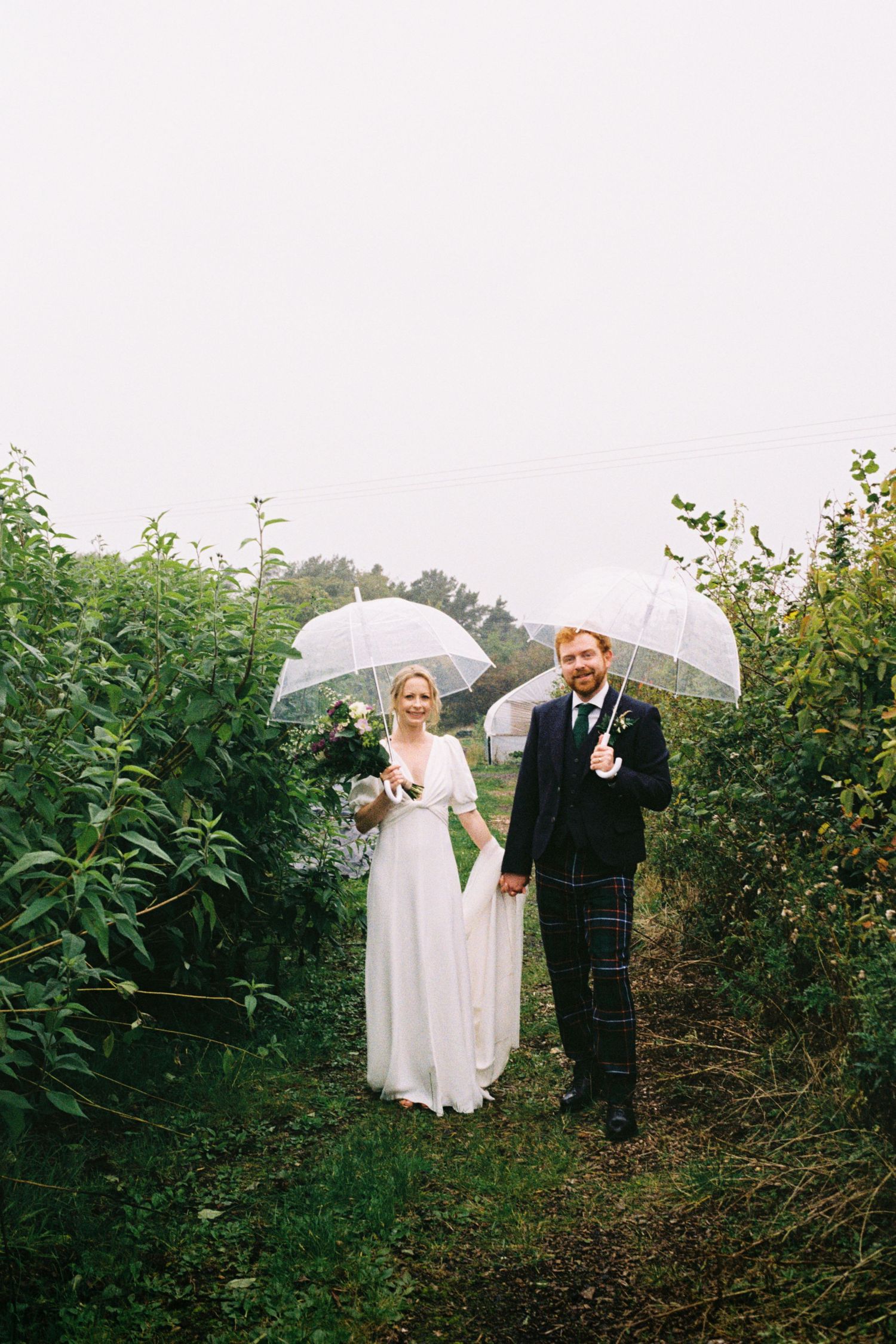 A couple holds white umbrellas while walking through an apple orchard on a misty wedding day.