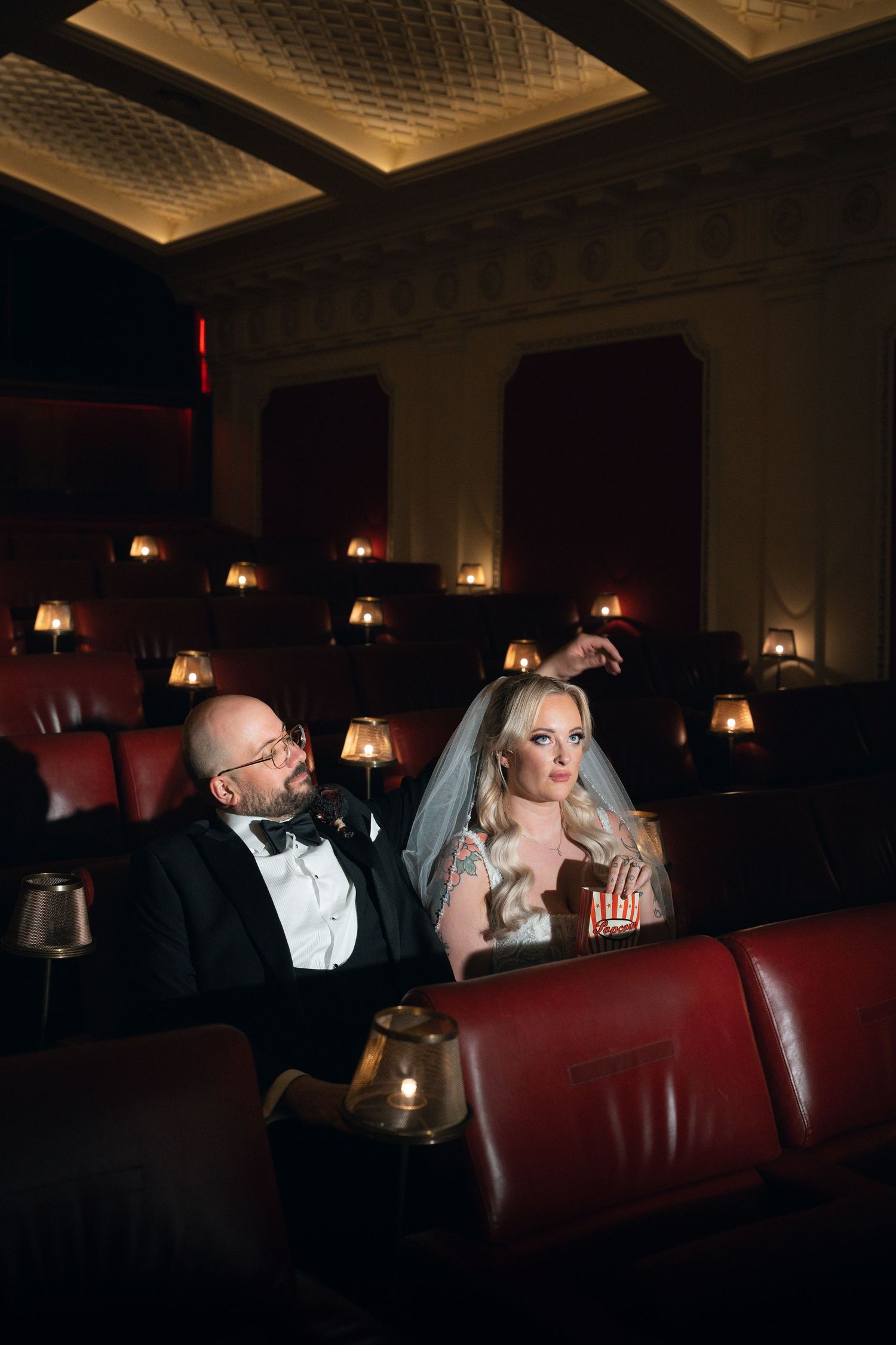 Bride and groom in wedding attire watching a movie in a luxurious vintage cinema with red leather seats and warm lighting.