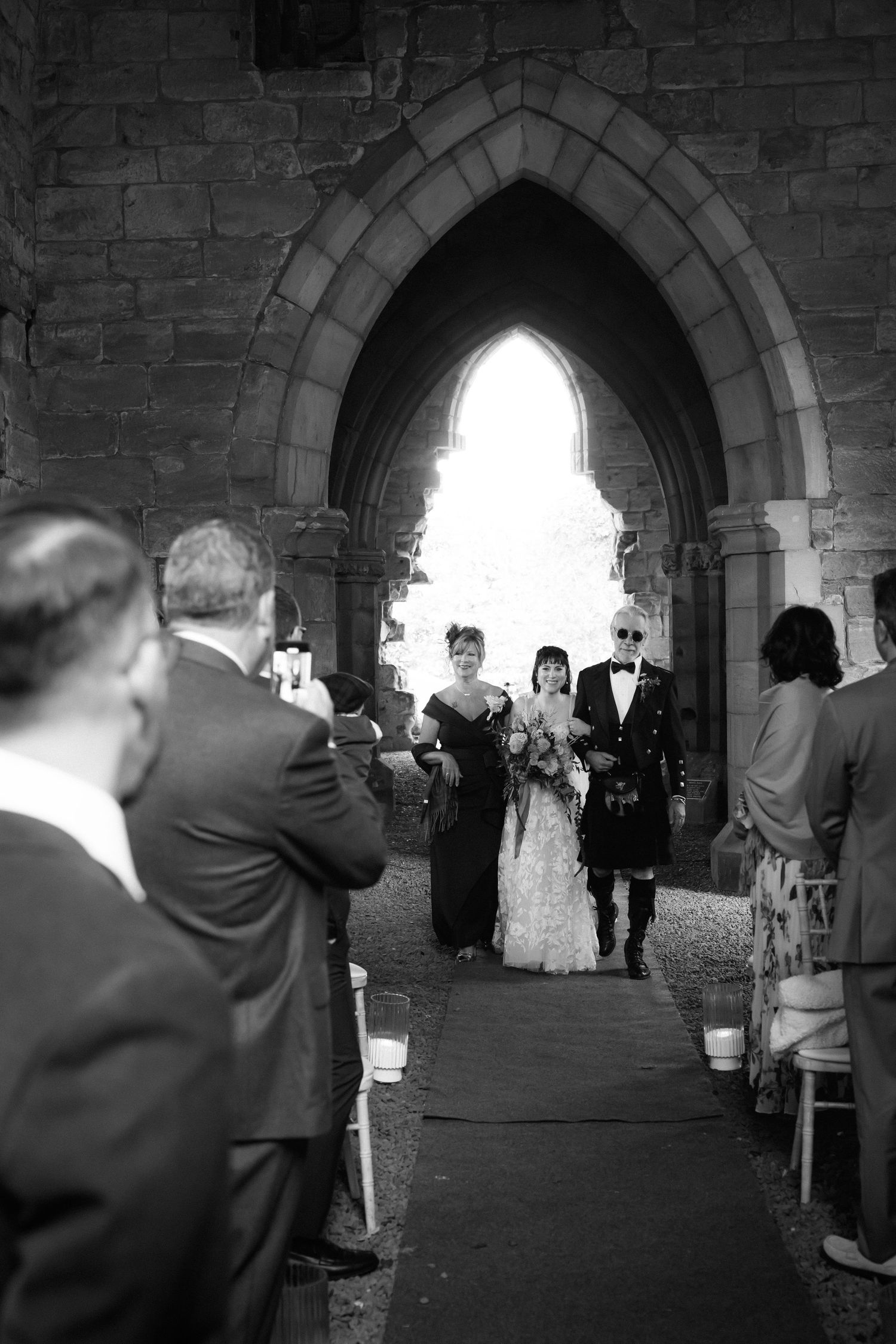 A black and white wedding ceremony photo showing a couple in an arched stone church doorway with guests looking on.