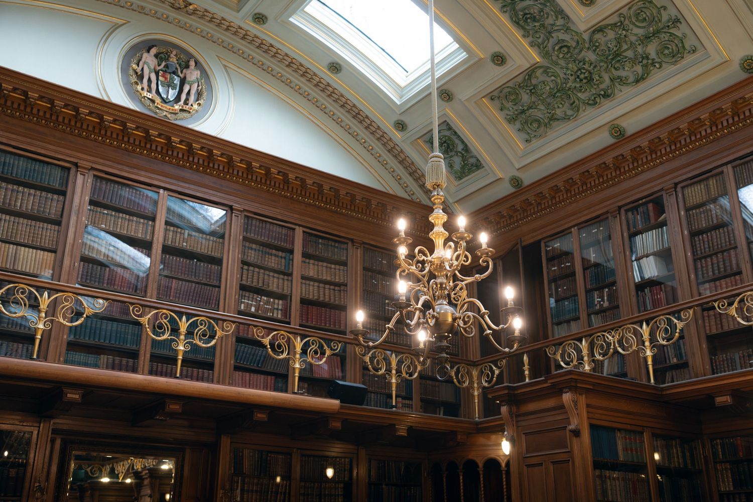 Elegant library interior with ornate ceiling, skylight, chandeliers, and wood-paneled walls filled with books.