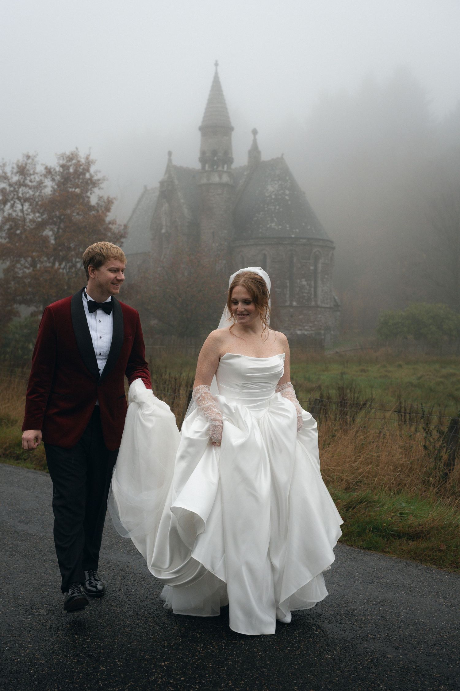 A couple walking through fog in front of a gothic castle on their wedding day.