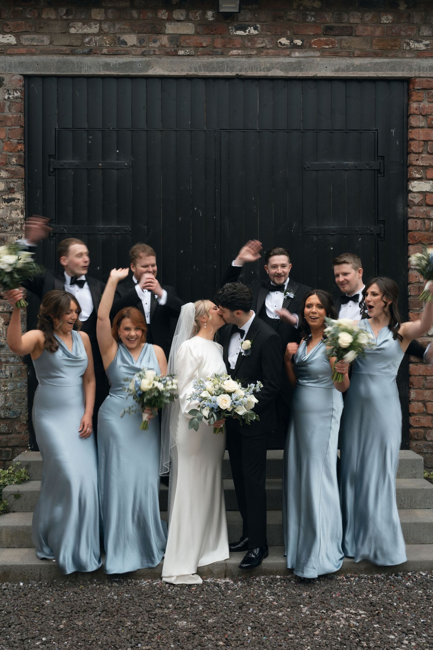 Wedding party in dusty blue gowns and black tuxedos celebrate as bride and groom kiss before rustic brick wall.