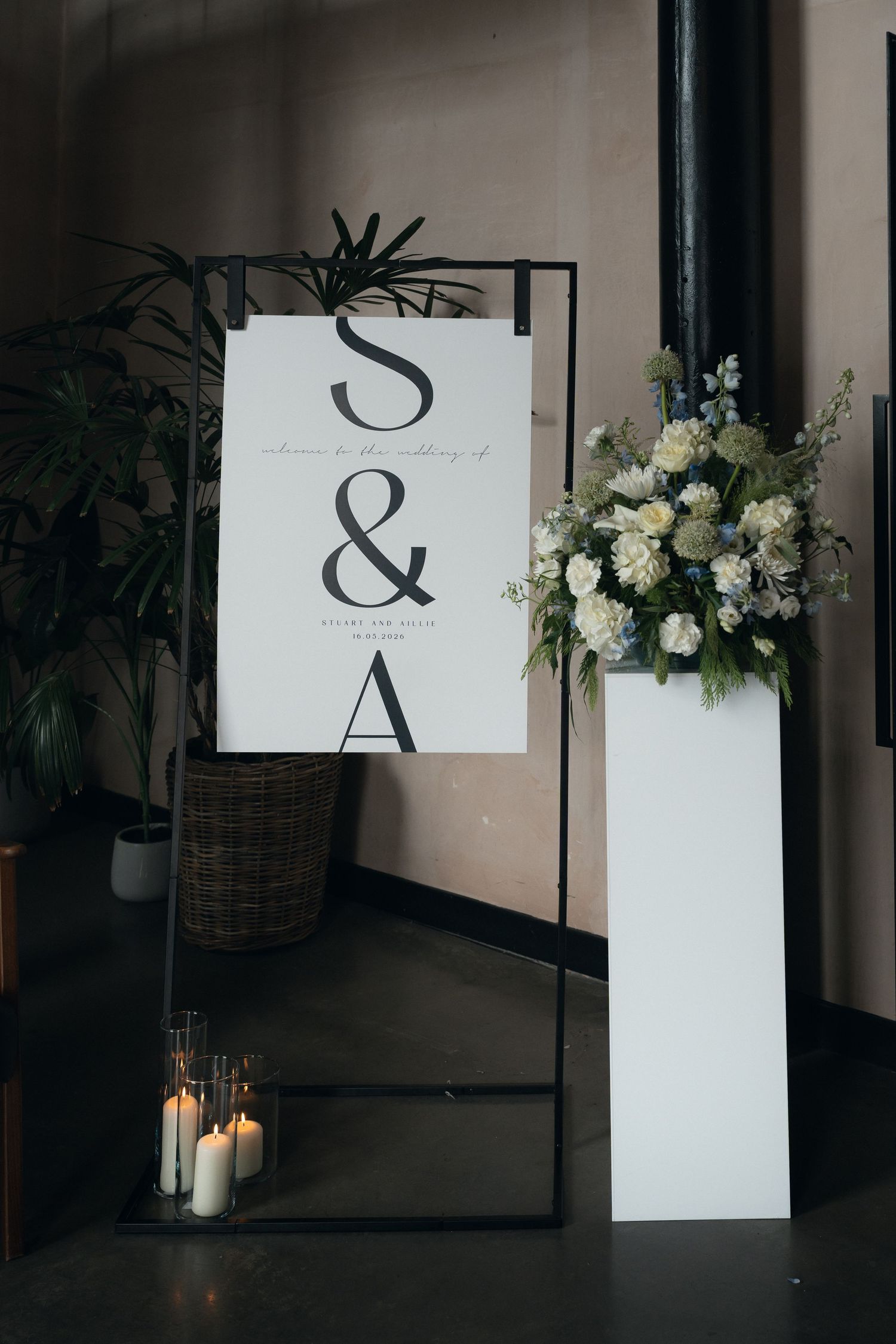 Elegant wedding welcome sign with S&A monogram on black metal stand, flanked by white floral arrangement and candles.