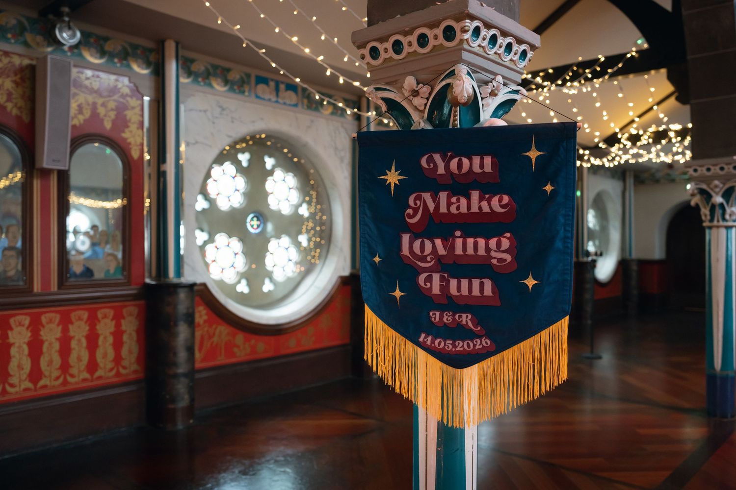 Decorative banner reading You Make Loving Fun with gold fringe in a colorful fairground interior with string lights.