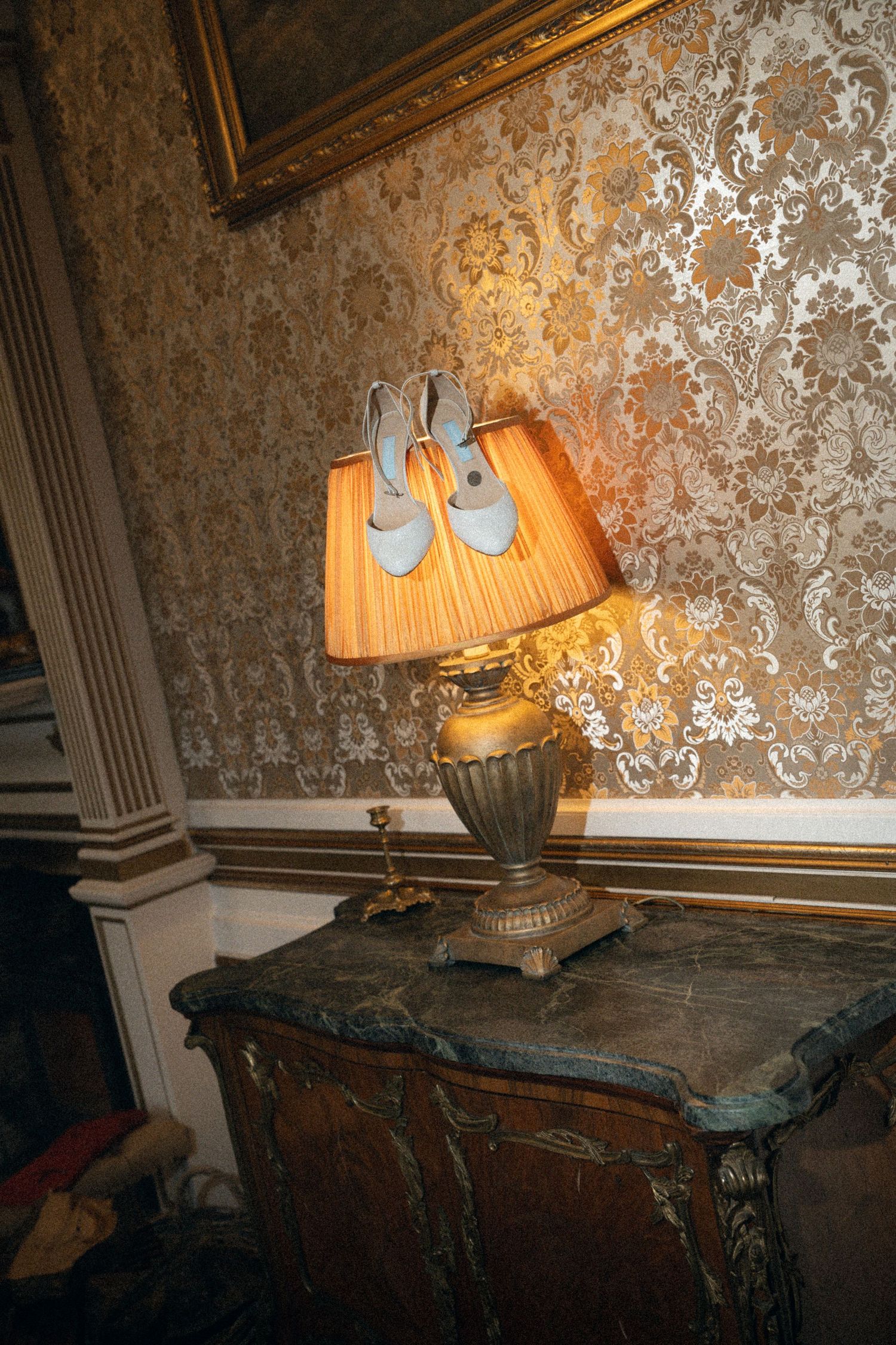 A vintage table lamp with pleated shade sits atop an antique dresser against ornate wallpaper.