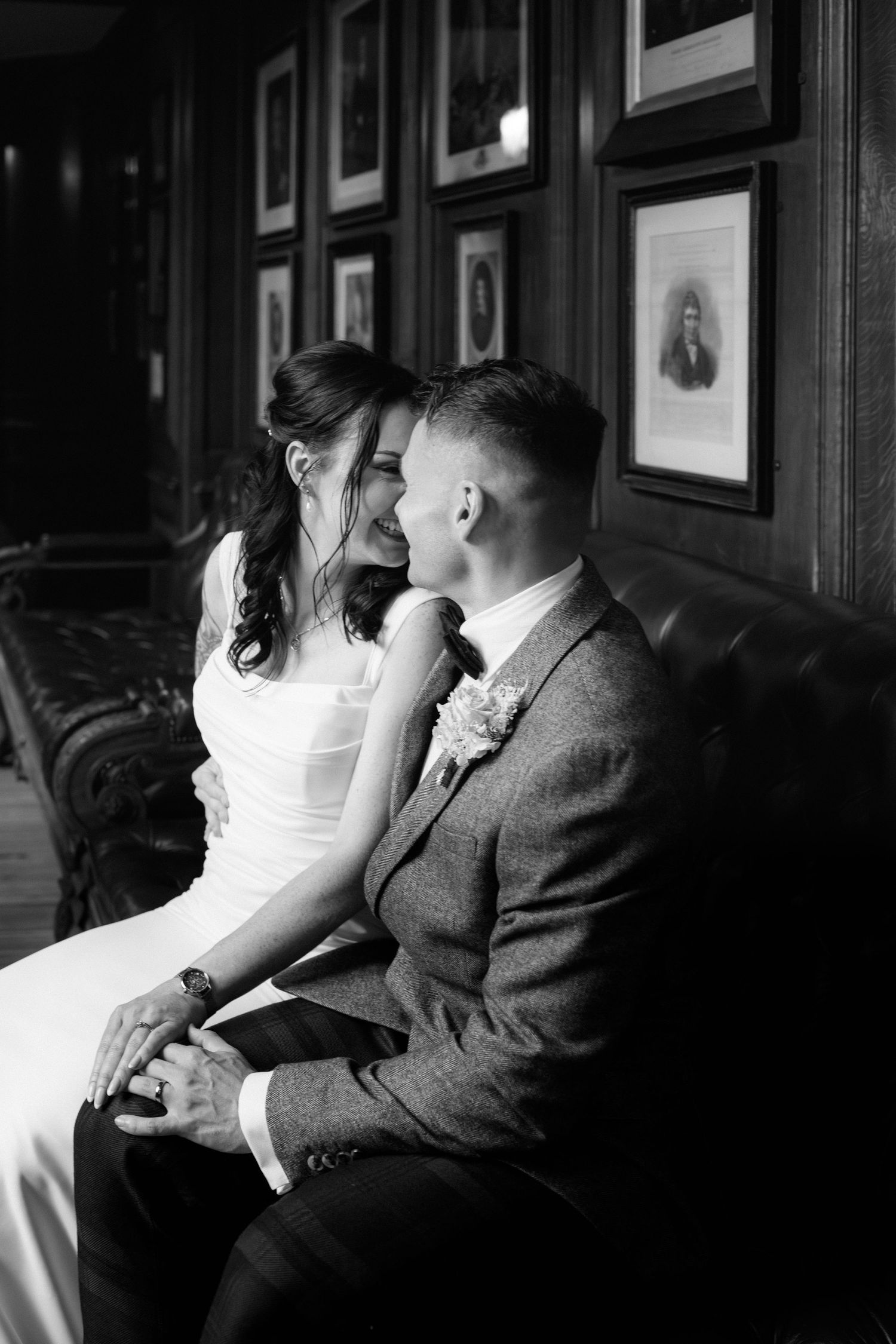 A bride and groom share an intimate moment on a leather couch with vintage portraits hanging on the wall behind them.